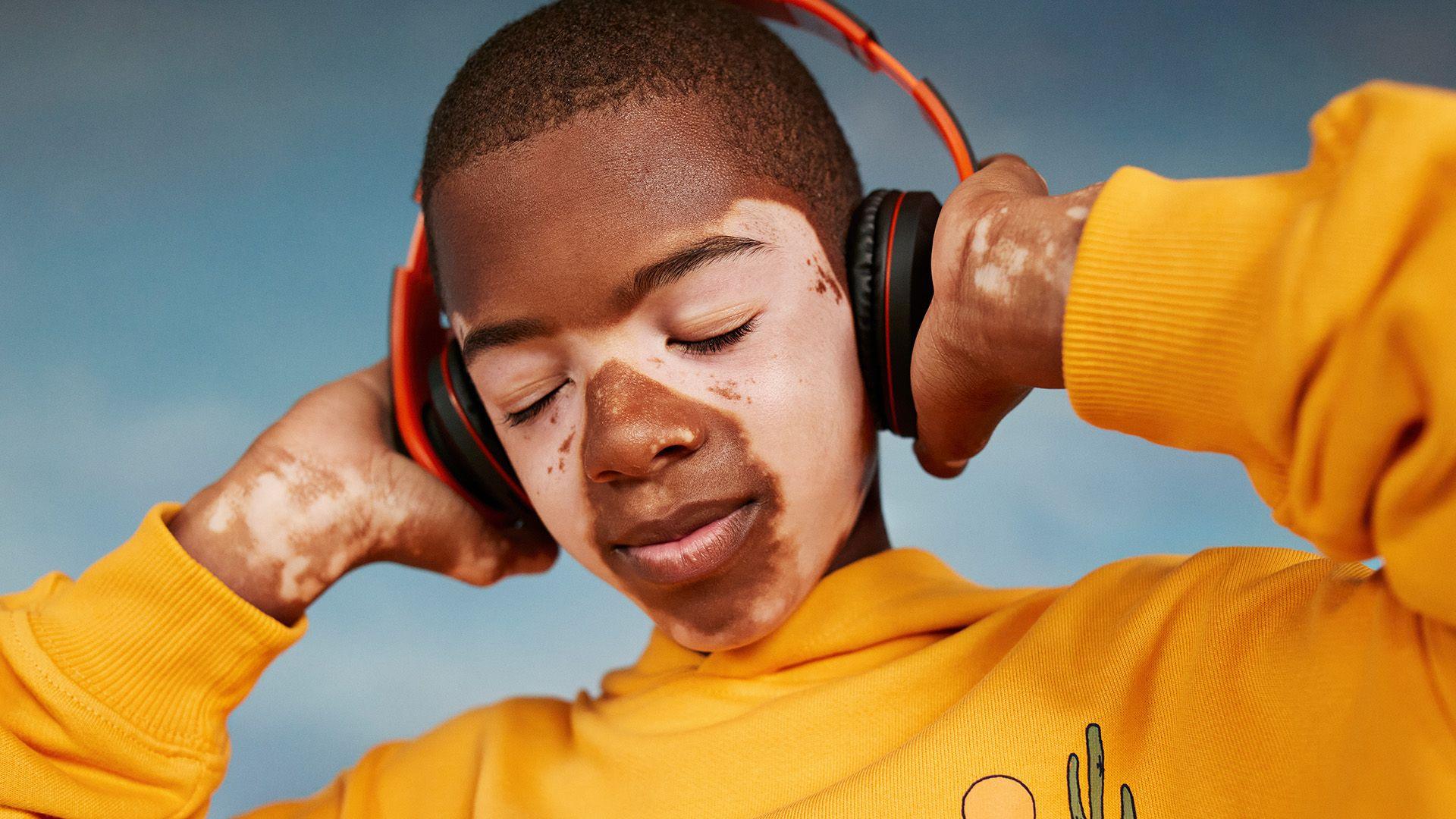 A boy with a yellow hoody on wears black and red headphones. He has is eyes closed and is holding the headphones over his ears
