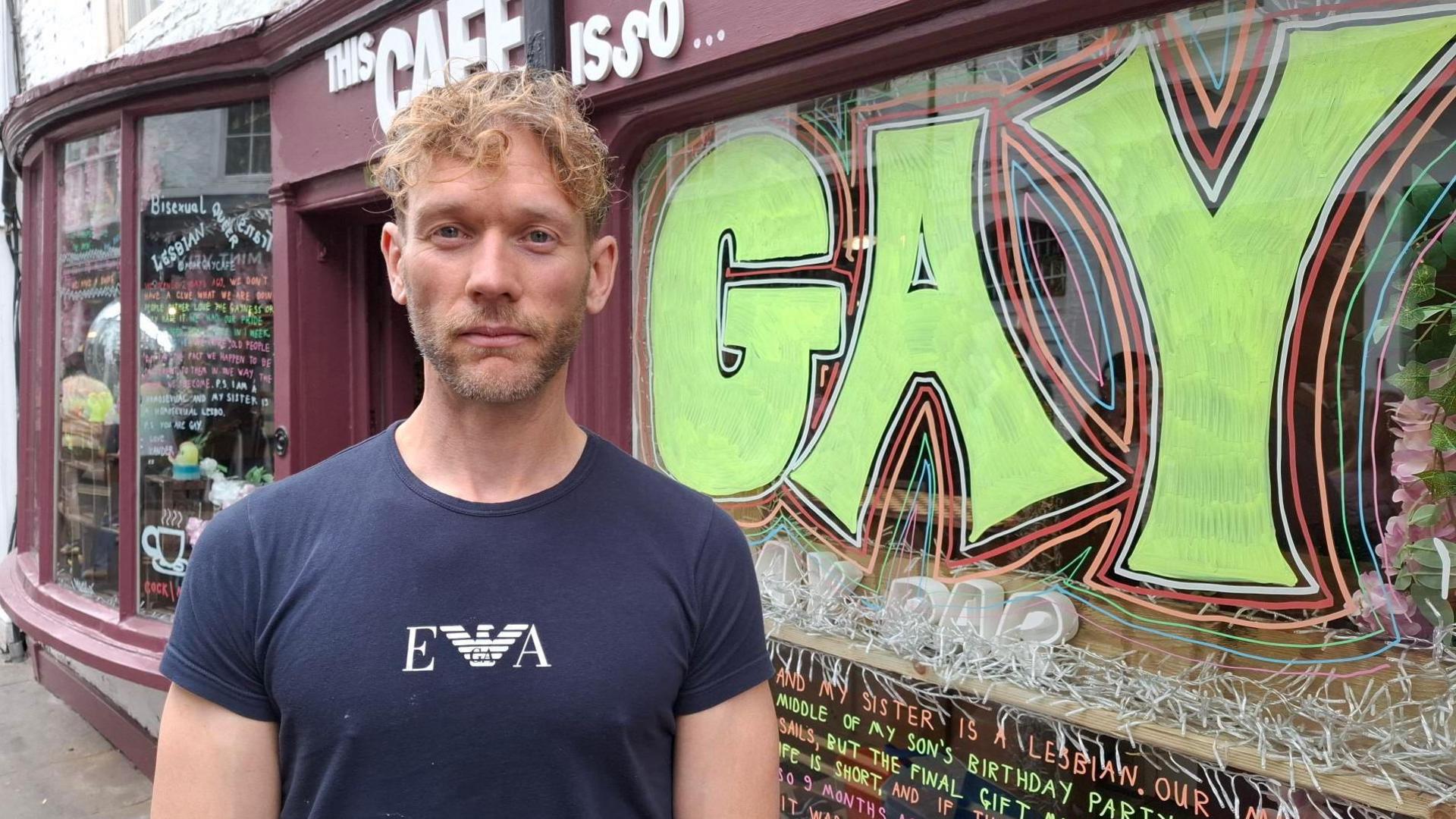 A man wearing a navy T-shirt with a logo on it. He has short blond wavy hair and a short beard. He is standing outside a shop, which reads "this cafe is so gay", with the final word in huge brightly-coloured lettering.