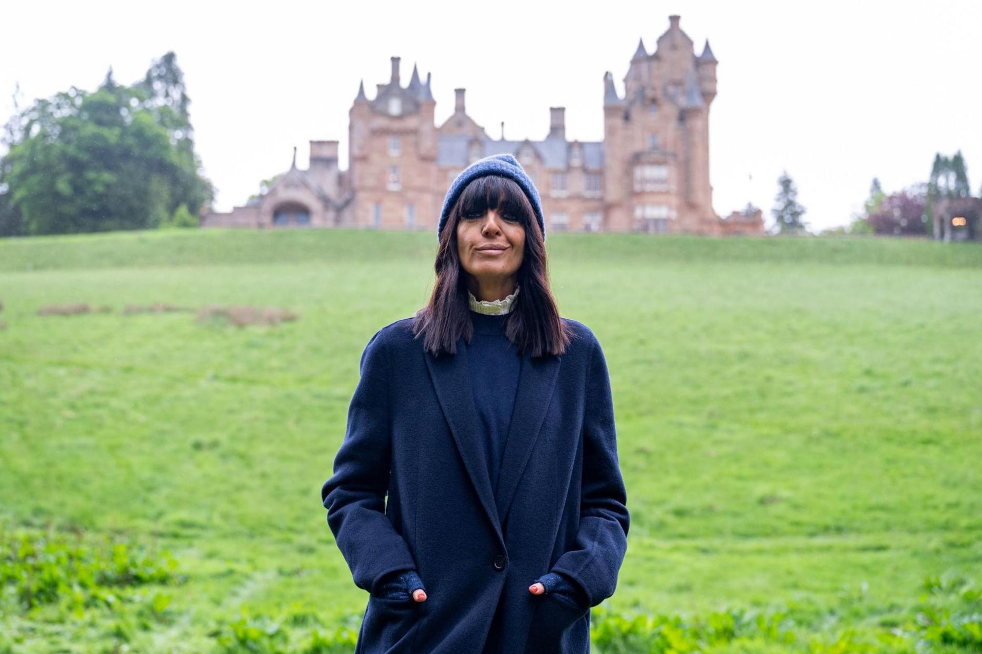 BBC host Claudia Winkleman is wearing a blue jacket and blue woolly hat while she stands at the bottom of a grassy slope below the towers and turrets of Ardross Castle.