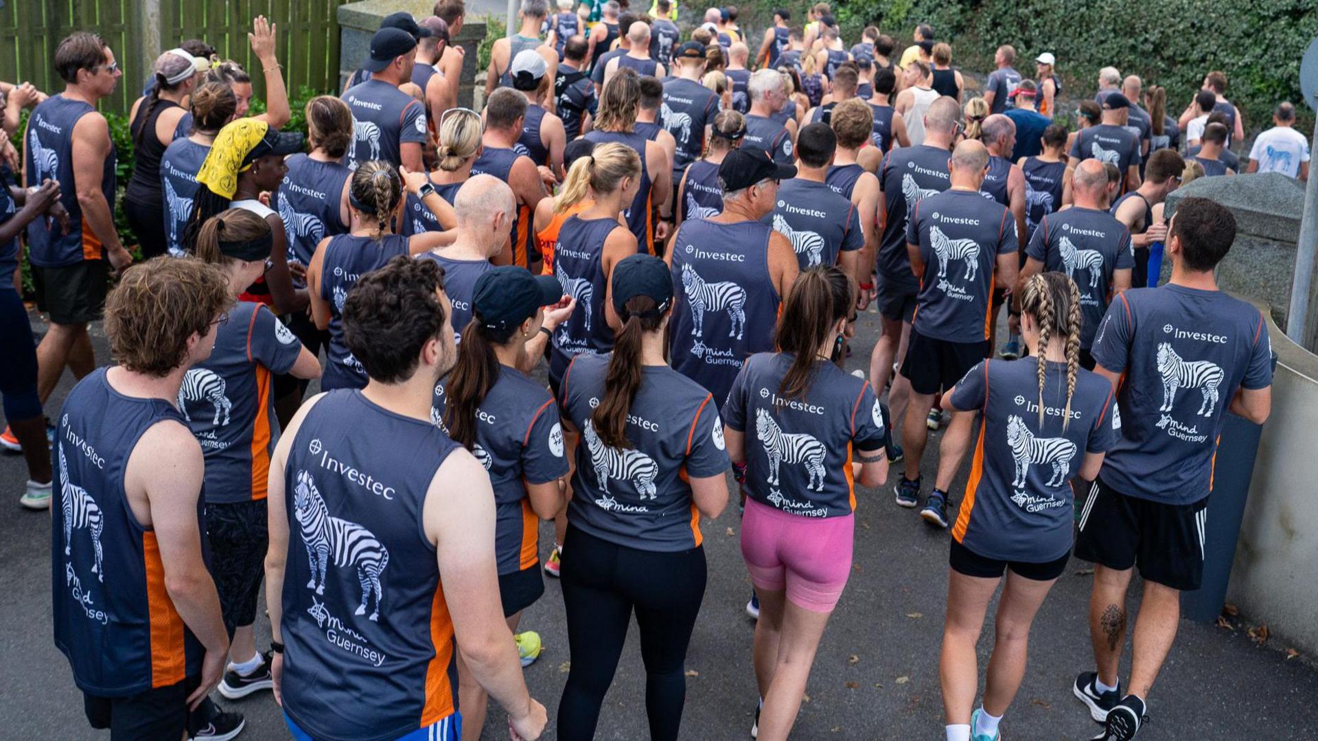 Image shows part of a crowd of hundreds of people in running-gear gathering for the start of a previous 10K Challenge to raise funds for Mind Guernsey. Most of the runners are wearing dark grey T-shirts supporting the charity, while a girl in the foreground is wearing pink shorts. The runners have their backs to the camera.