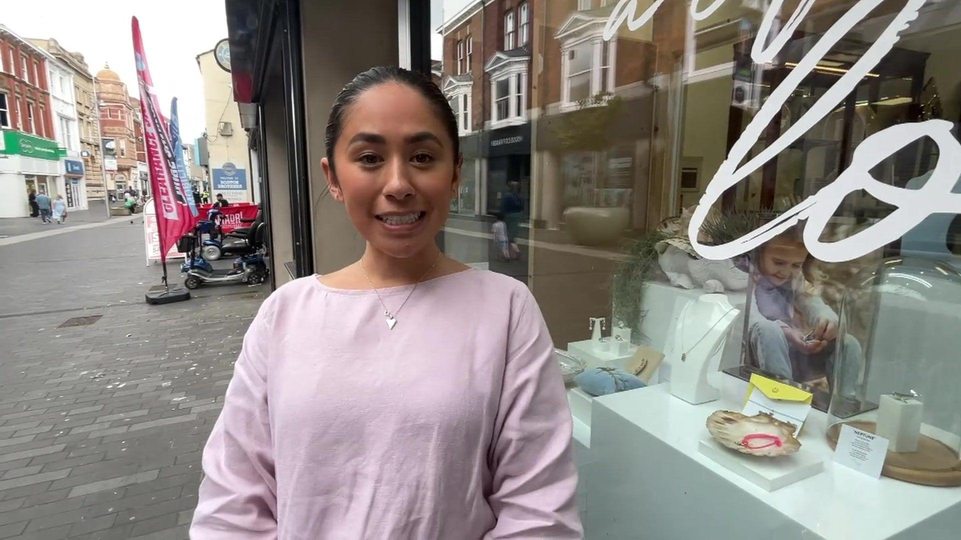 Brianna Yeung stands outside of the jewellery shop where she works. She is wearing a lilac t shirt and a silver necklace with a heart on it.