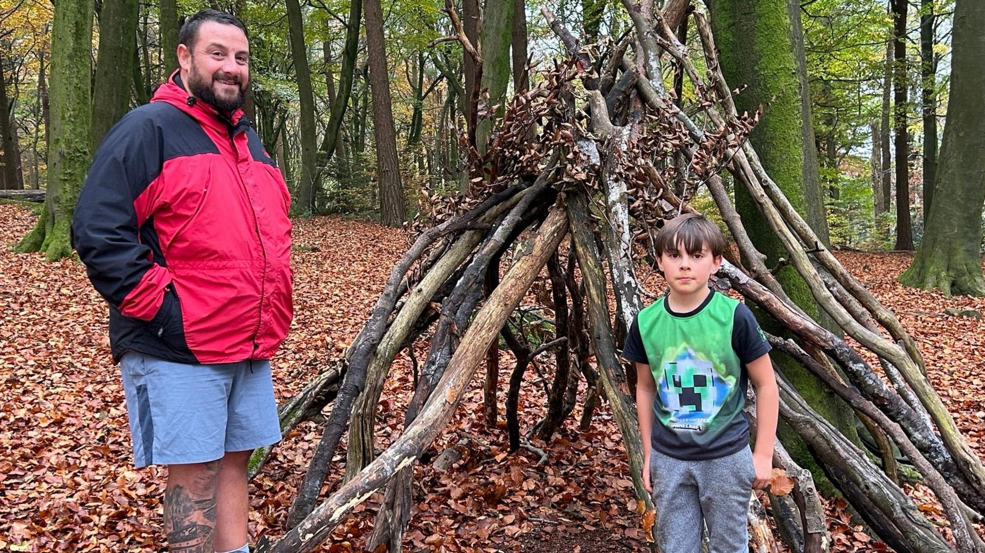 Samuel, wearing a green Minecraft t-shirt and grey jogging bottoms, stands in front of a den constructed using large branches in a woodland area. He is watched by a smiling member of staff from an independent school which provides specialist support for autistic children.