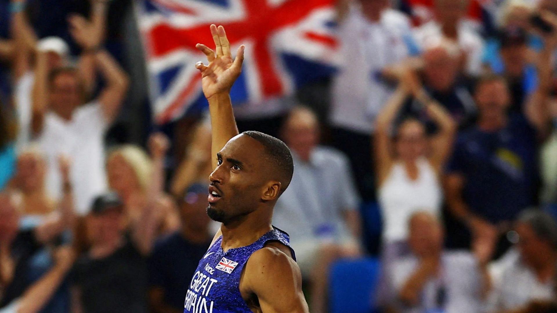 A man wearing a blue vest holding up a hand in front of a crowd and a Union Jack flag Great Britain's Matthew Hudson-Smith celebrates after winning the gold medal in the Men's 400m.