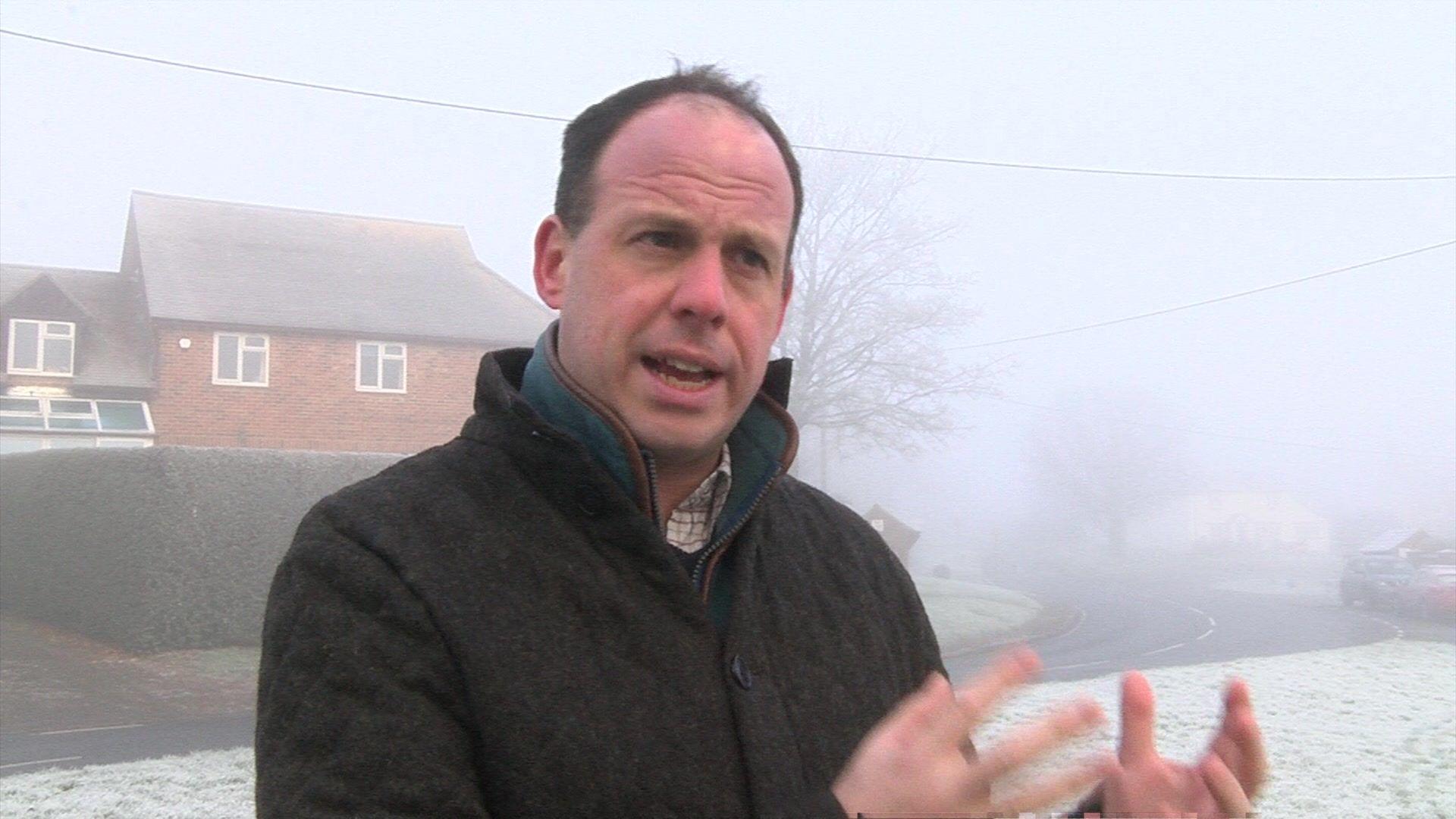 MP Greg Smith gestures with his hands to express his feelings against the biogas plant. He stands on a grass verge near a road, with a house and hedge behind him, on a very foggy day. He wears a white checked shirt, a teal jumper and a cushioned black jacket.