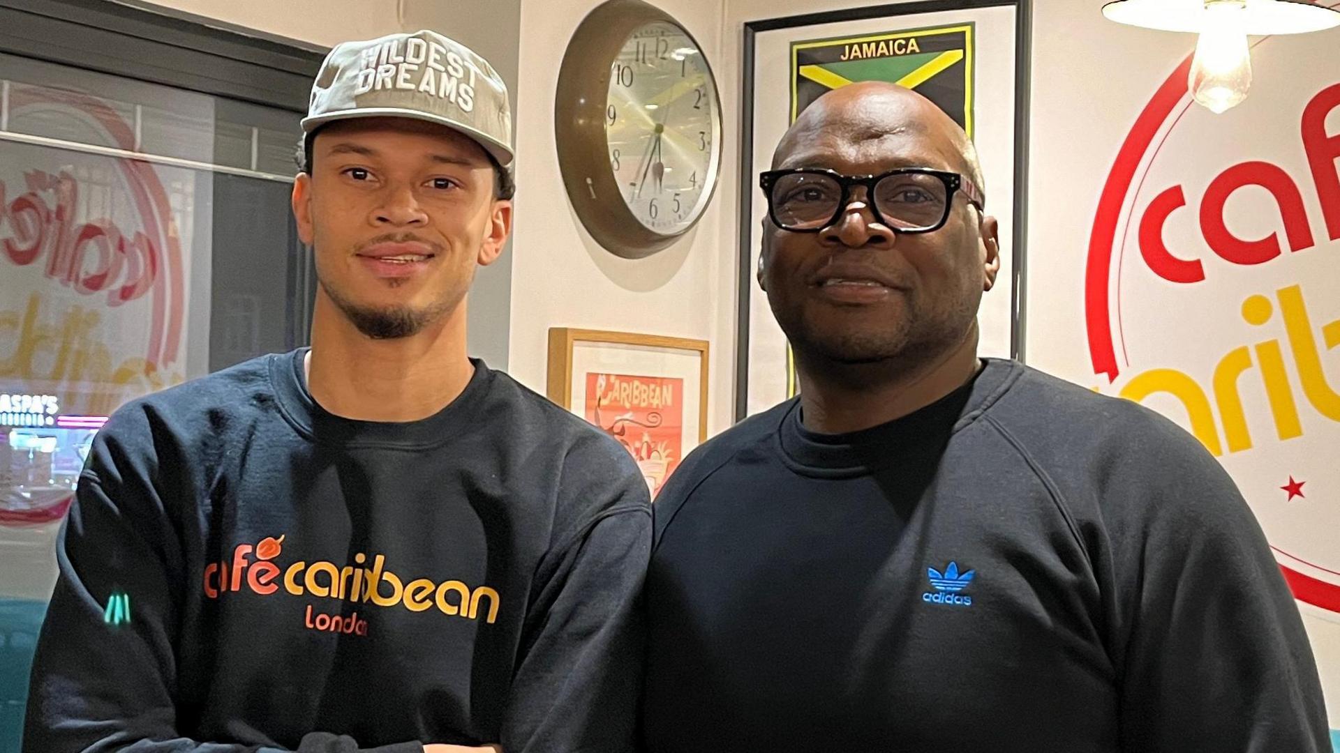 Warren Richards and one of his sons inside the cafe. Mr Richards is wearing black glasses and a black Adidas jumper while smiling at the camera. His son is wearing a black jumper that has "Cafe Caribbean" printed on it in yellow and red font. He is also wearing a light green cap that has "wildest dreams" on it.