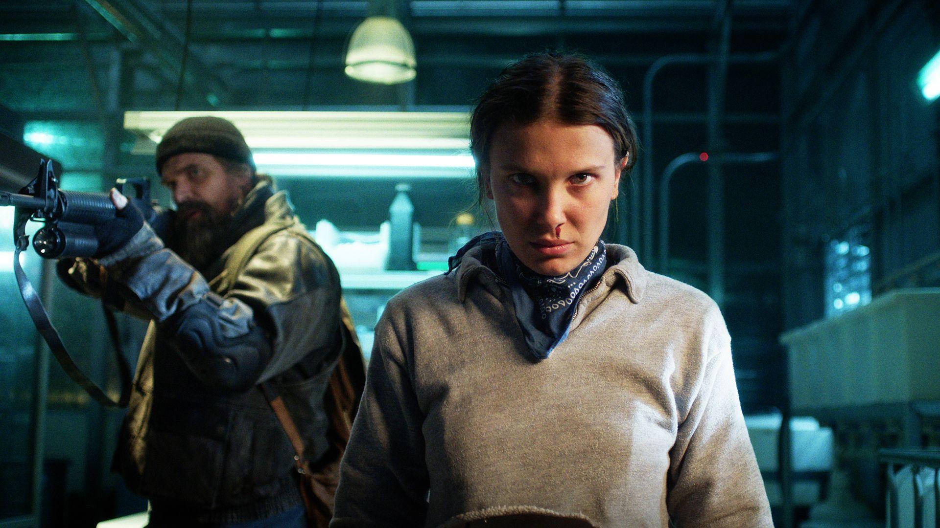 (L to R) David Harbour as Jim Hopper holding a gun and Millie Bobby Brown as Eleven looking straight at camera in Stranger Things.