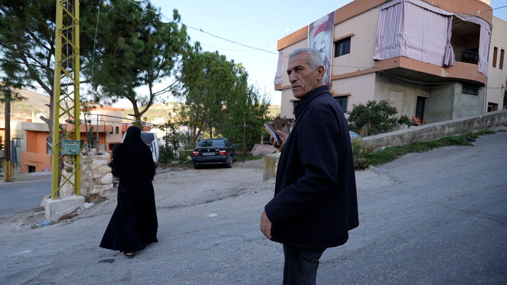 An older man is seen on a road, with a woman nearby, in front of a building which has a large image on the side.