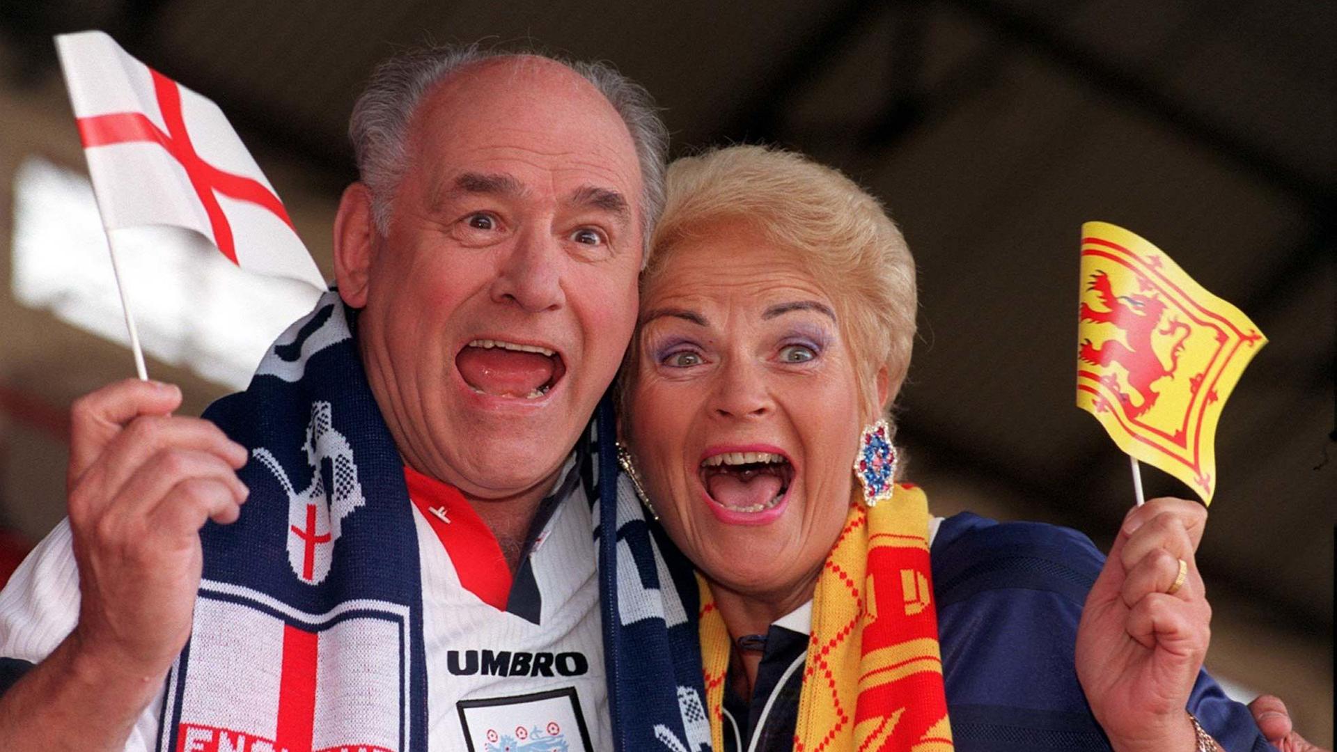 A man and a woman dressed up in football gear wave flags. They look happy and excited.