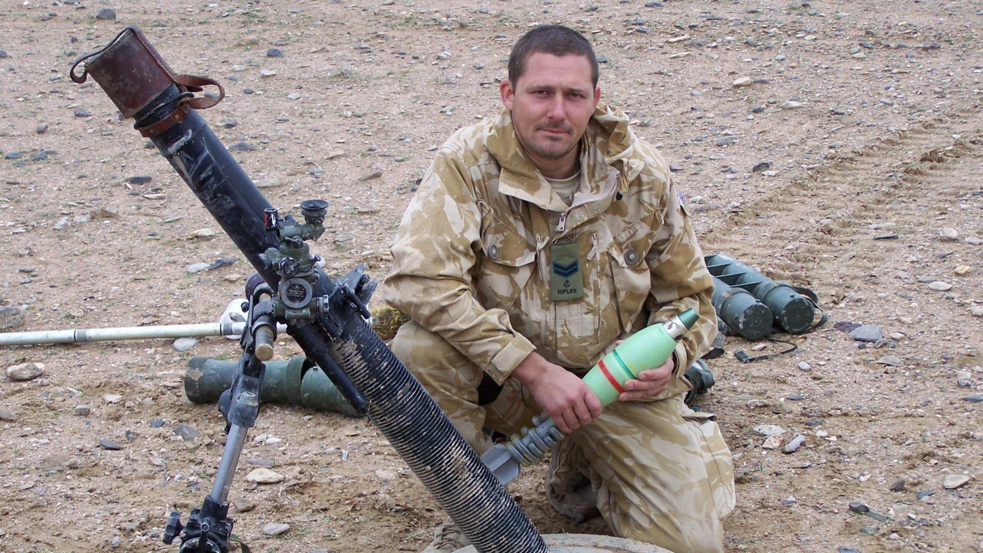George Pas wearing beige and cream camouflage uniform and kneeling down in the dirt beside a shoulder fired missile launcher.