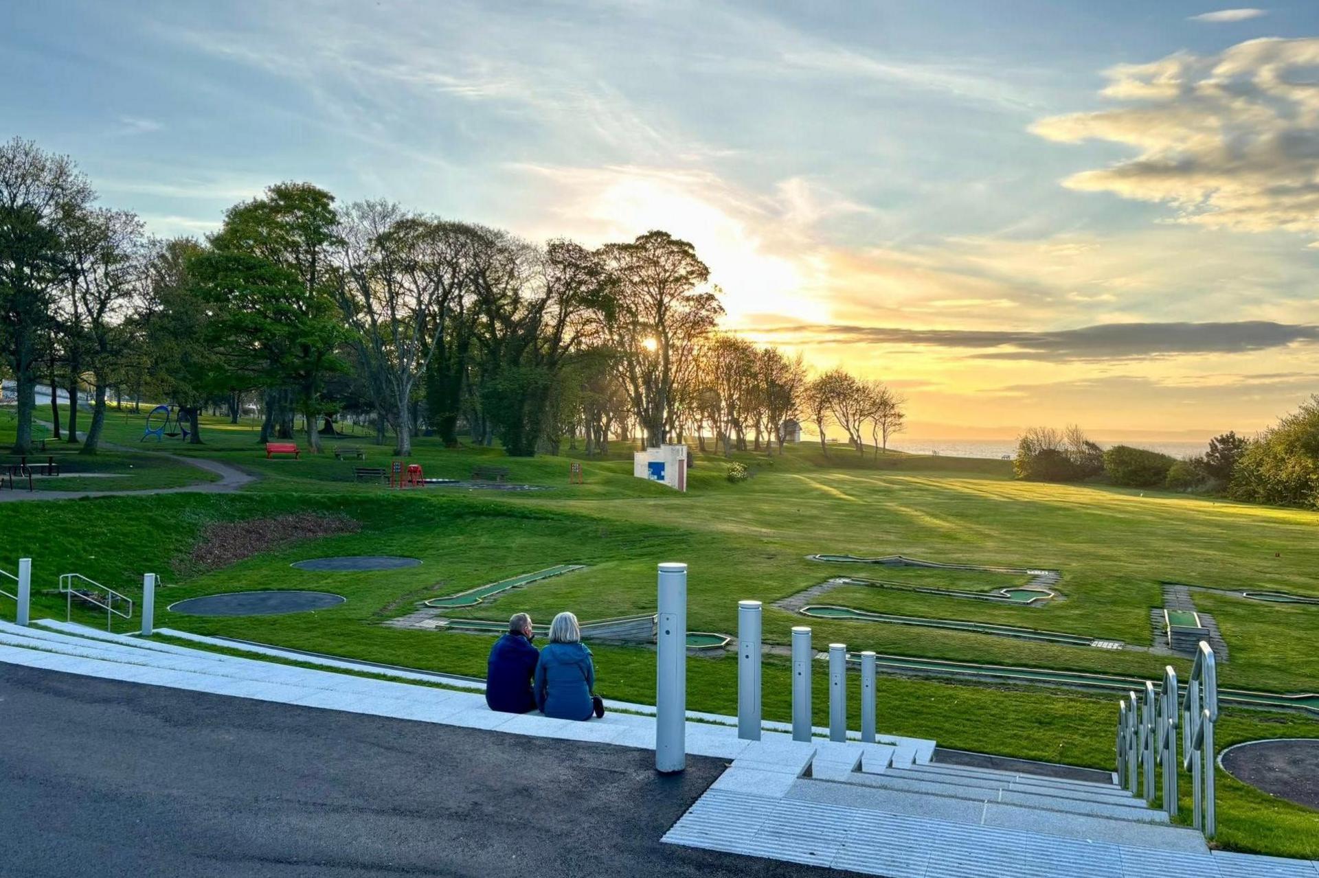 Two people sit together on the terraced seating and look out of a gently rolling area of parkland towards the sea.