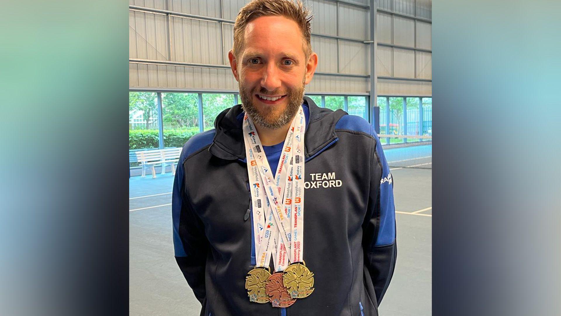 James Lawton has short brown hair, glasses and brown stubble. He is wearing a jacket that says Team Oxford on it and has three medals around his neck. They appear to be two gold medals and one bronze medal.