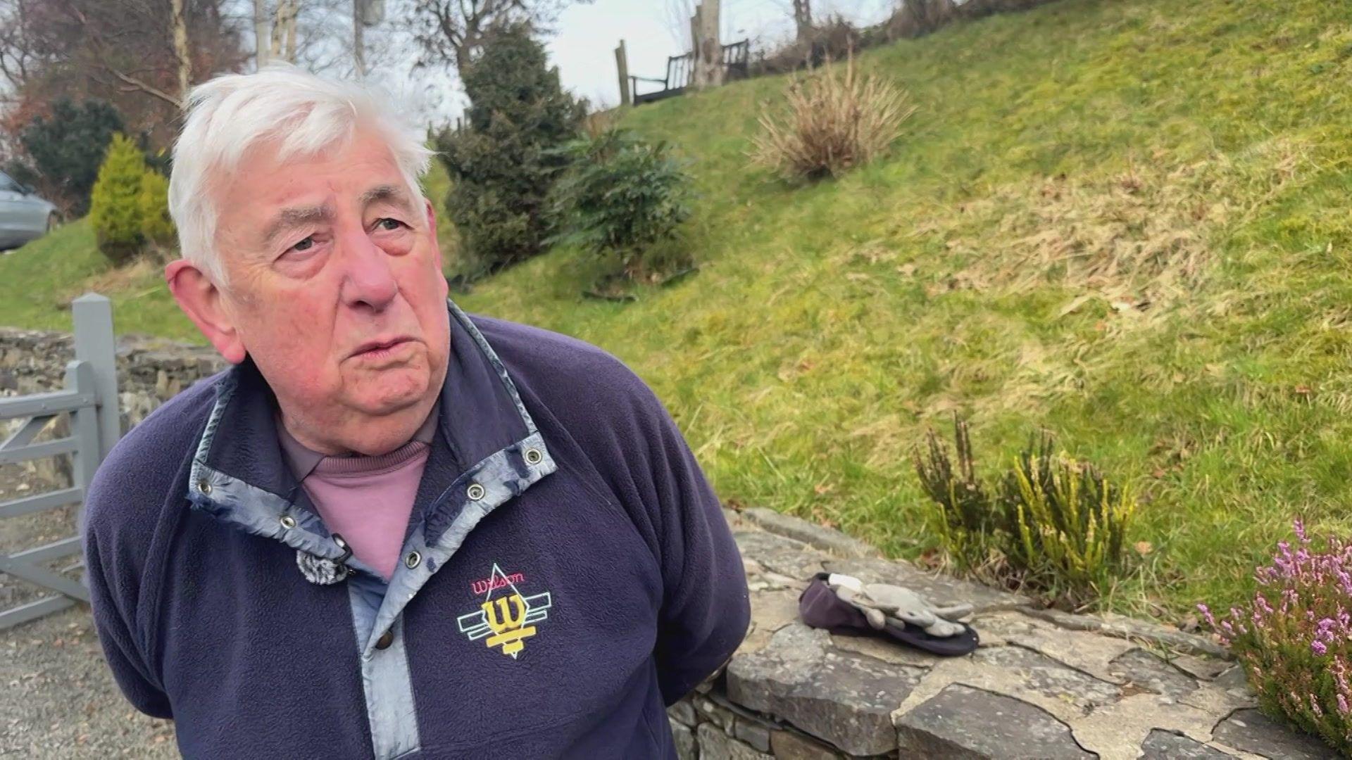 Tony Littlewood, has silver hair and wears a navy green half zip fleece. He is stood to the left of the image with a grey gate behind him and a grass hill next to a dry stone wall.