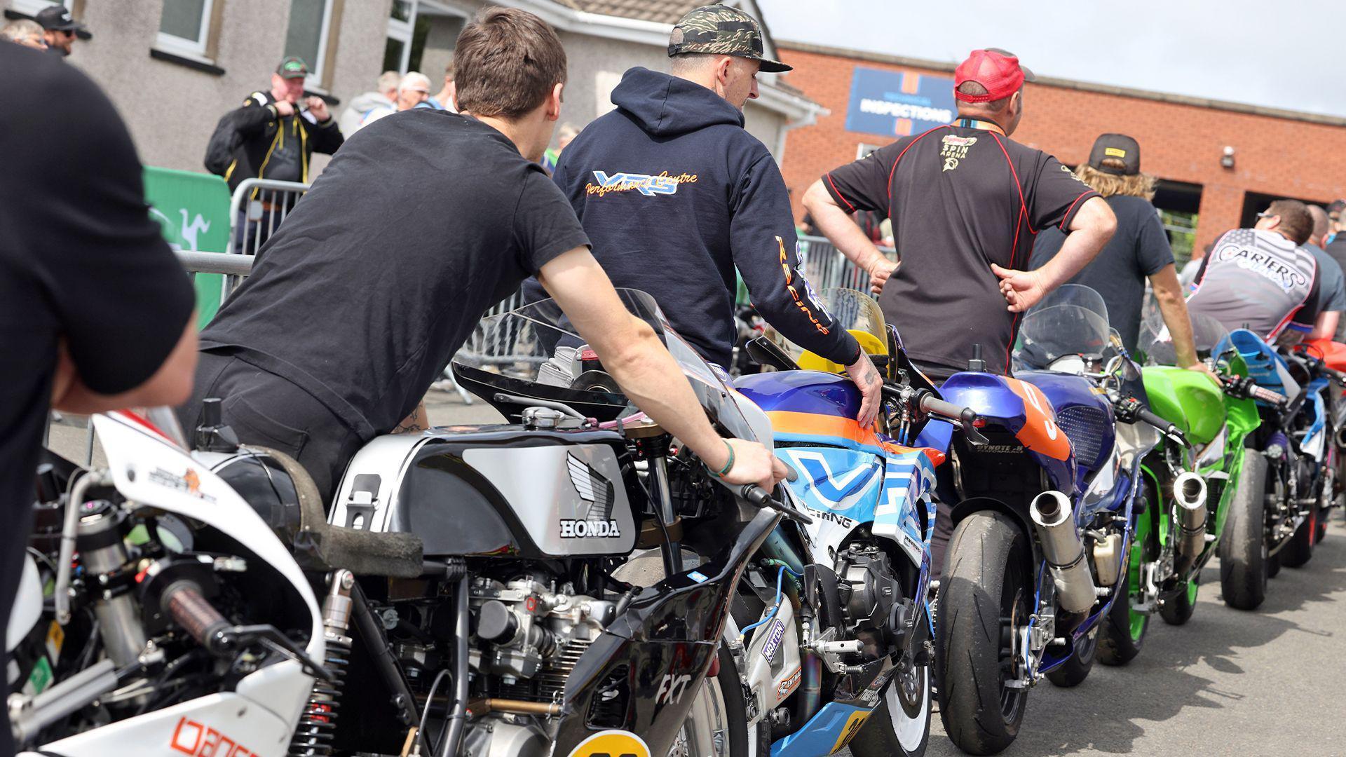 A row of motorcycles being wheeled up to the scrutineering section at the grandstand.