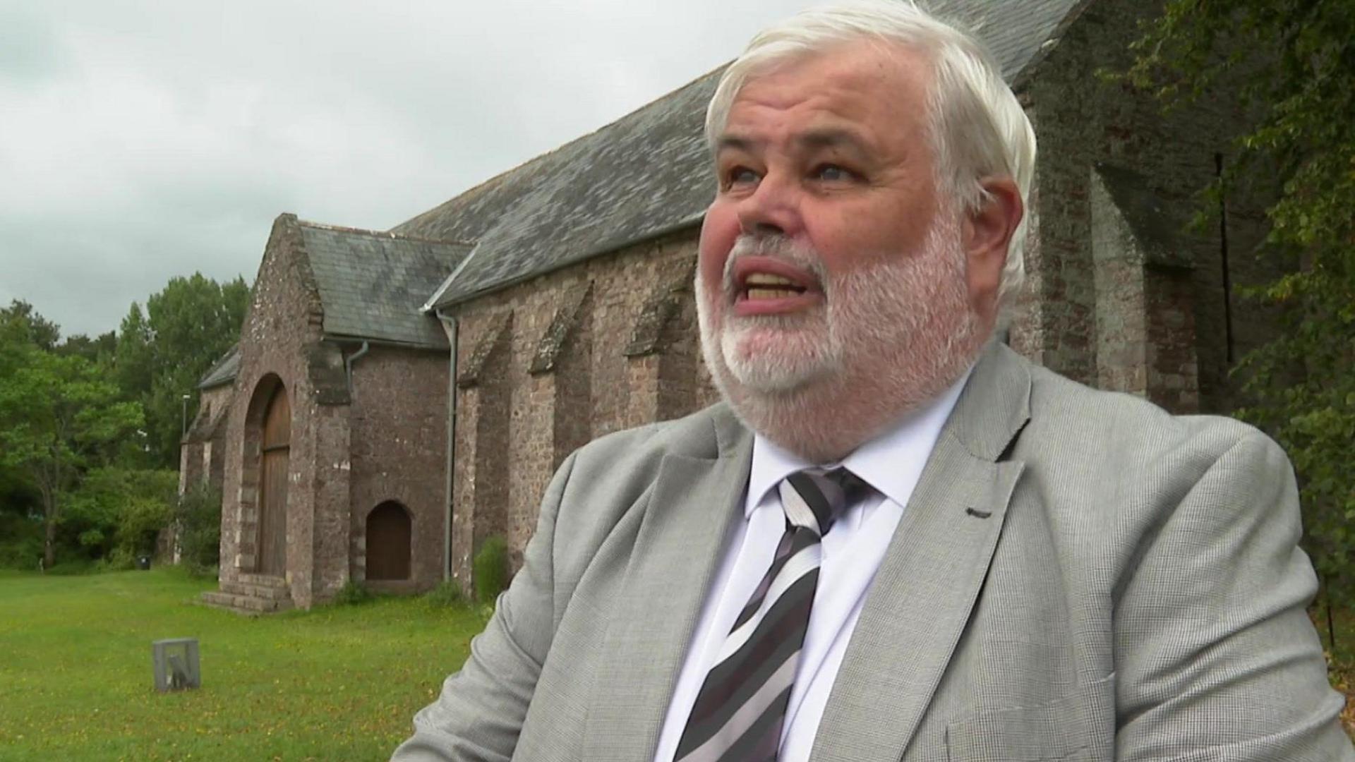 Torbay Council leader David Thomas stood in front of the Spanish Barn in Torquay. Mr Thomas has white hair and a white beard and is wearing a grey blazer, white shirt and a black, white and grey striped tie. He has his mouth slightly open mid-speech. The Spanish Barn is a windowless stone building with a large wooden door.