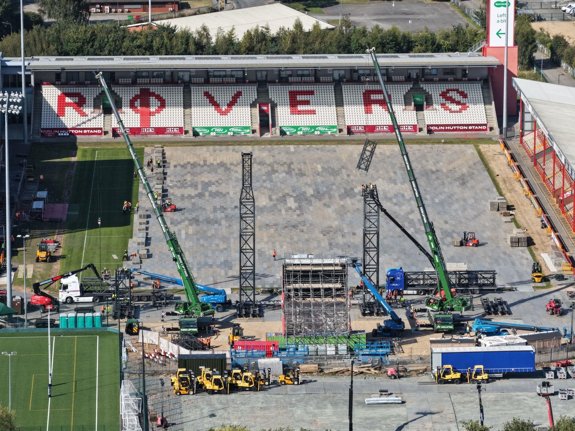 A construction site inside Hull's Craven Park stadium, with cranes and machinery on the field. The stand in the background displays the word "ROVERS" in red on white seats. Equipment and materials are scattered across the area.