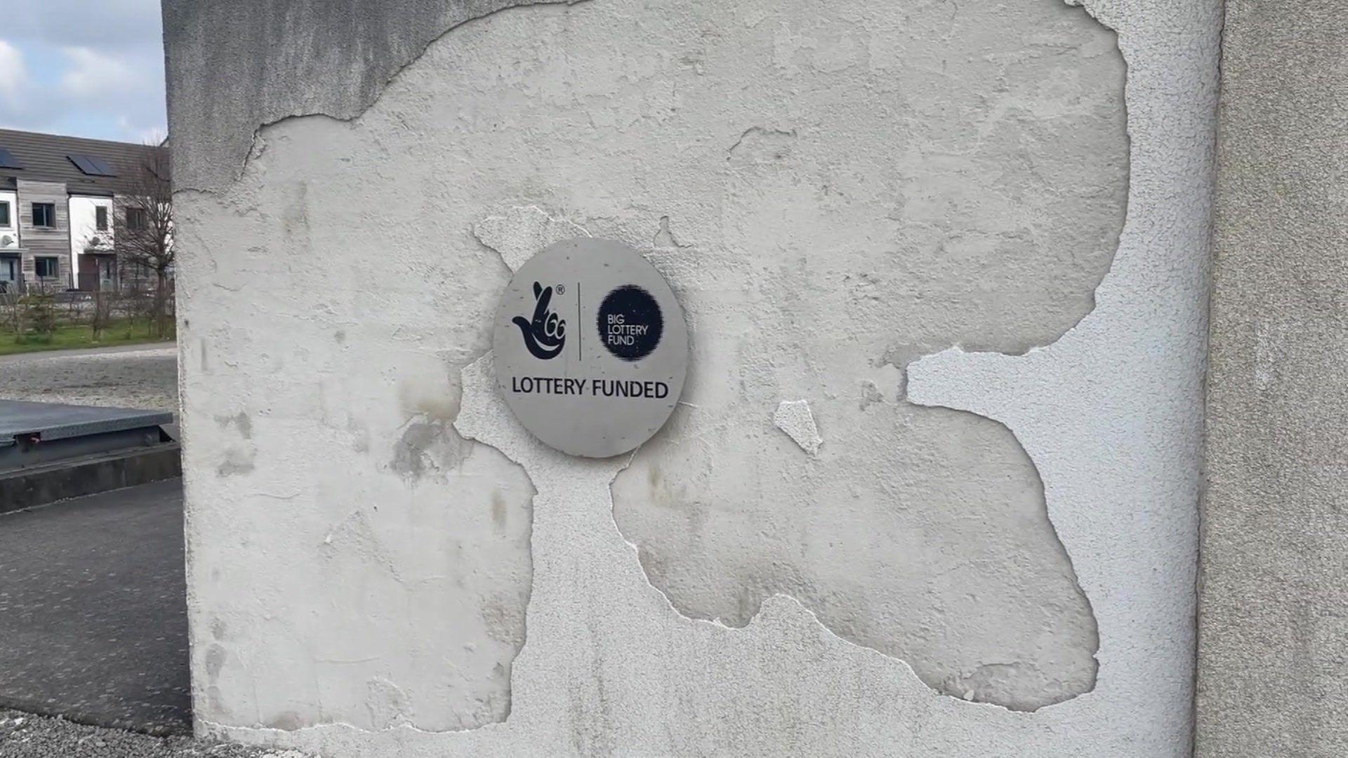 A small circular metal plaque on a wall saying: "Lottery funded". Concrete render is crumbling away around the plaque 