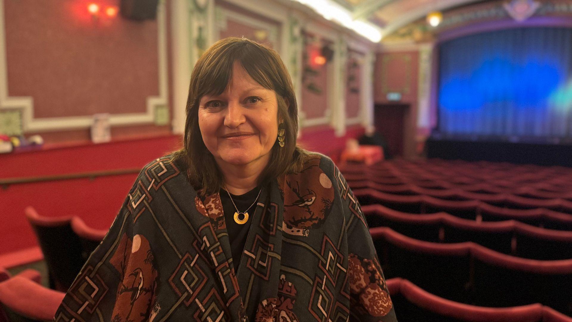 Deb Perkins is wearing a patterned top standing at the back of an empty cinema. The cinema's stage is in the background with a blue curtain in front of the screen.