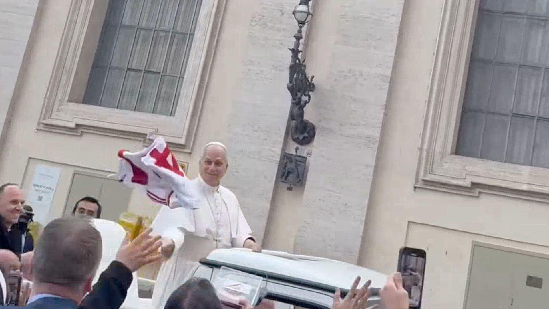 The jersey being thrown to Pope Leo. The red and white jersey is mid air and the Pope is smiling standing on the back of a truck.