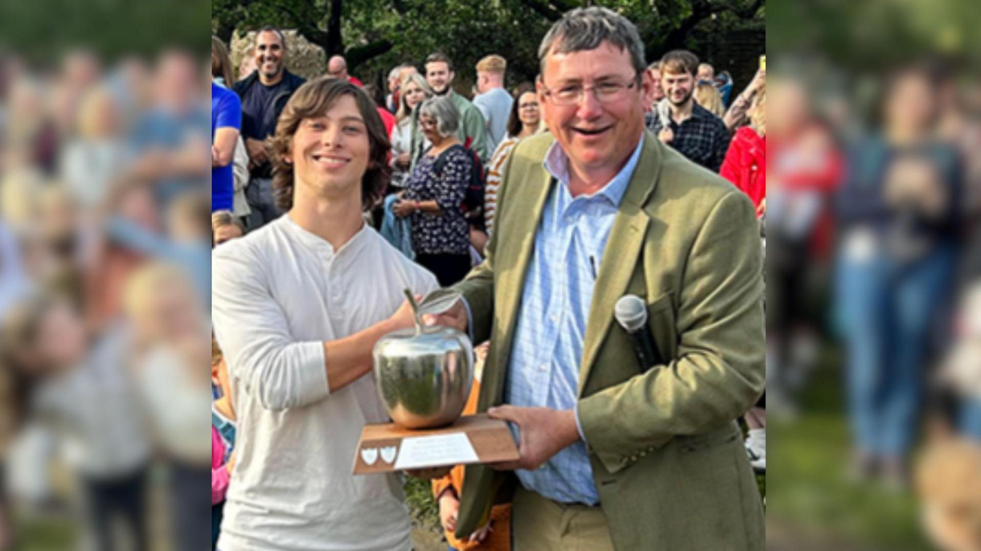 A smiling young man with long brown hair in a white shirt shaking hands with a older smiling man in a green suit jacket and blue shirt.

The man in the jacket is handing the other man an apple shaped chrome trophy and shaking his hand.

There are several people standing behind the pair of them