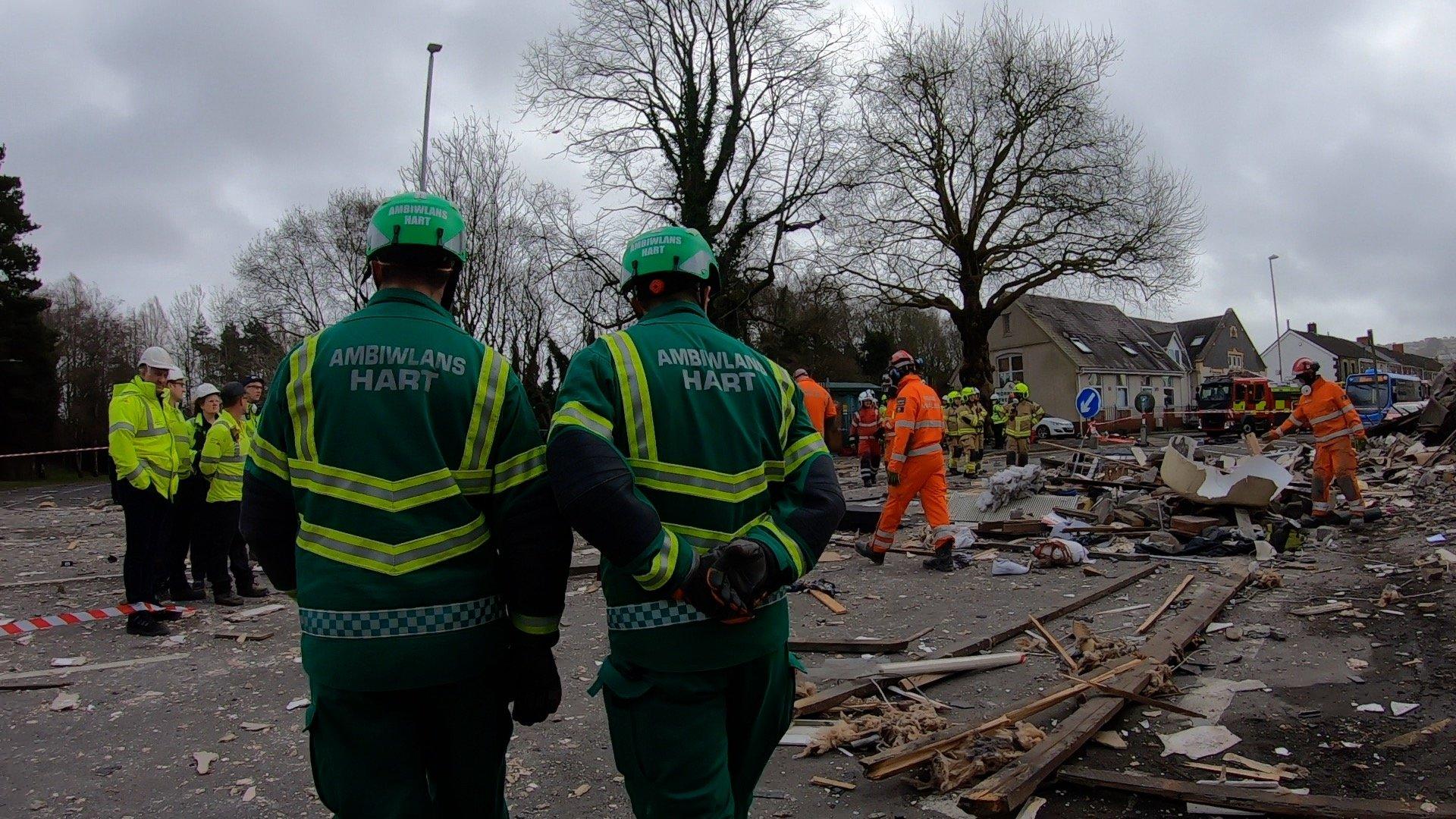 Two hazardous area response team members are at the scene of the explosion, alongside police officers and urban search and rescue officers. There is rubble strewn across the street.