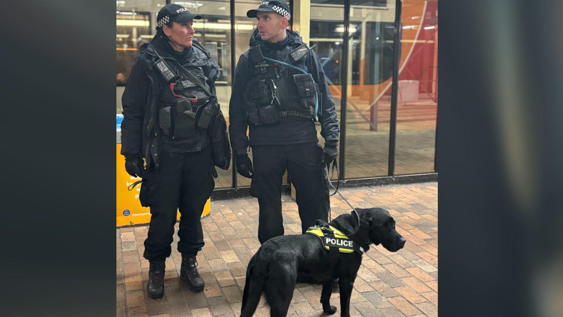 Police officers wearing black uniforms and black caps- standing in front of a black police dog with green jacket 