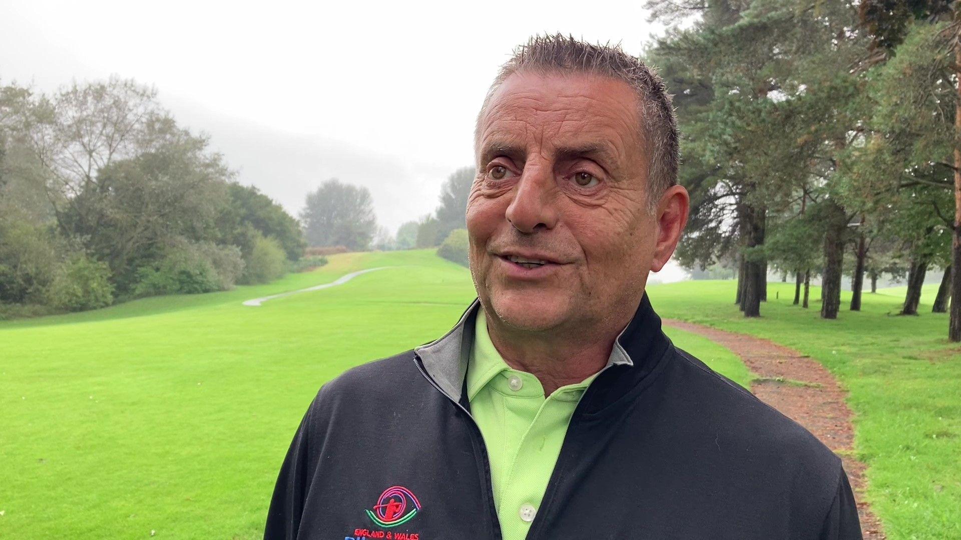 A man on a golf course, wearing a light green shirt and black golfing jacket. The gold course is lined with trees.