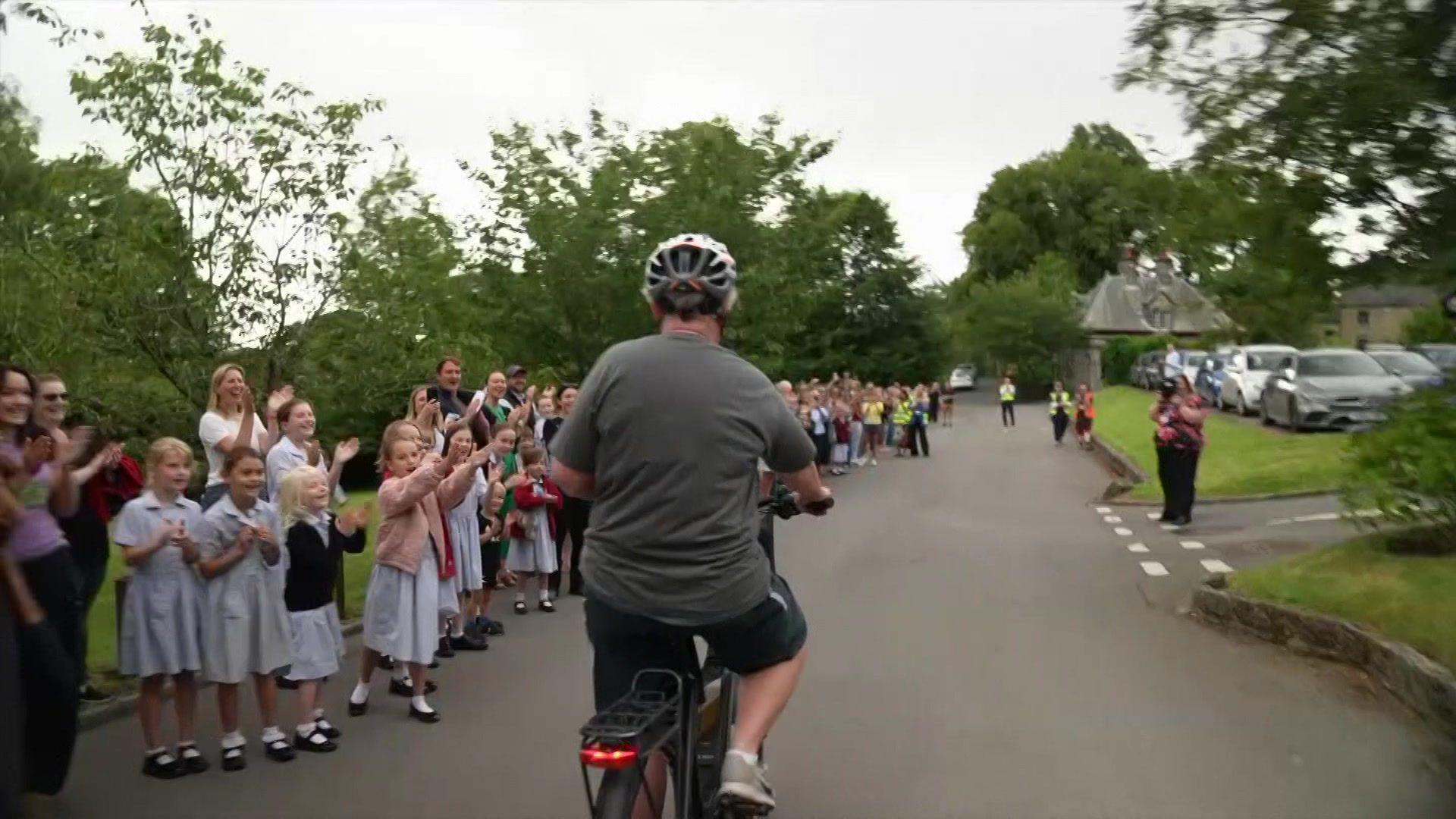 A line of children and parents is along the side of a long driveway, with a woman cycling down the middle, away from the camera.