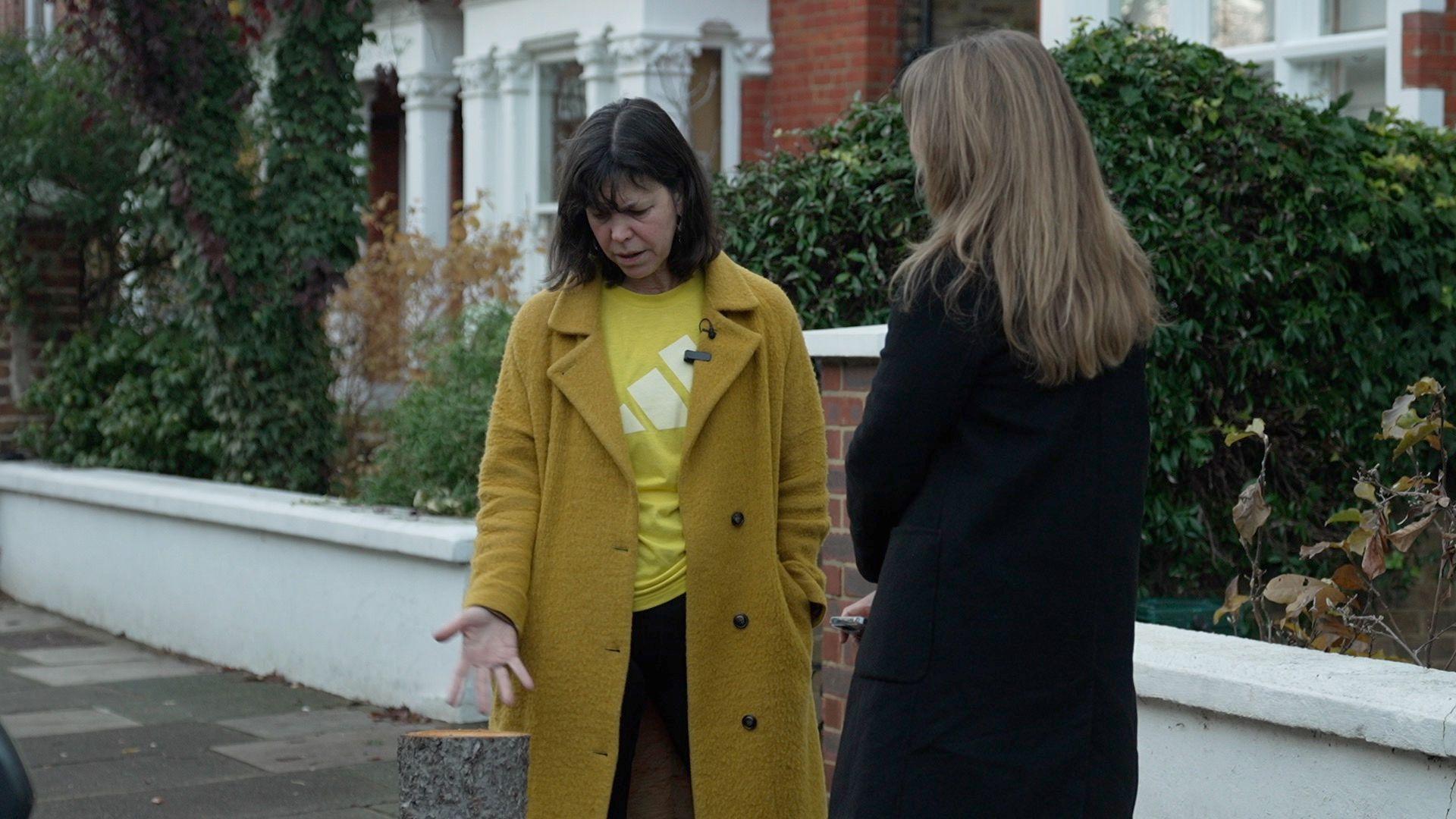 Alison Murphy is wearing a yellow coat and gestures at the stump of the tree while speaking to a reporter in a black coat. A white wall is in the background.