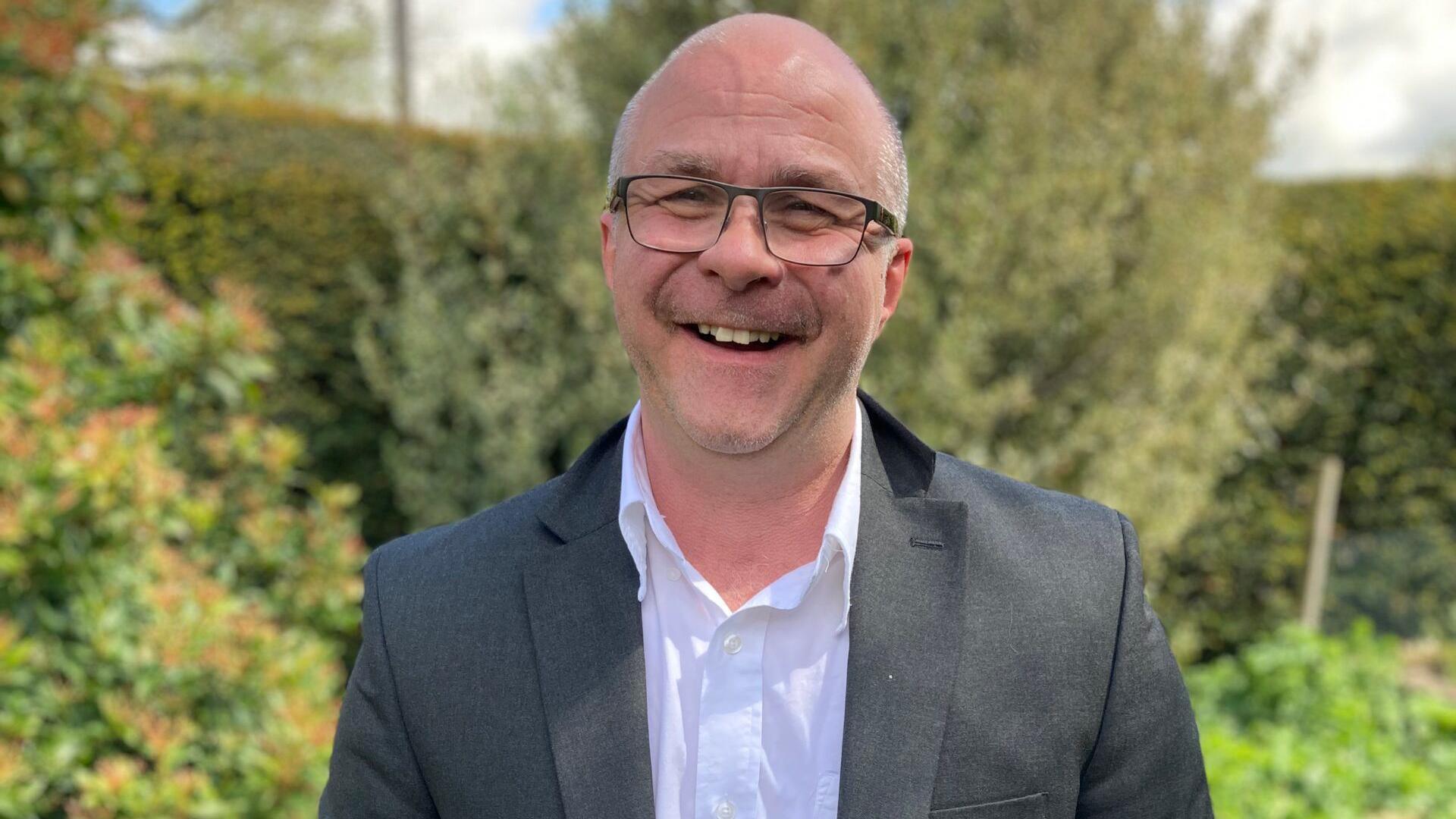 A man with very short white hair and black glasses is smiling at the camera. He has a white shirt on with a black suit blazer. Behind him are green bushes and trees. 