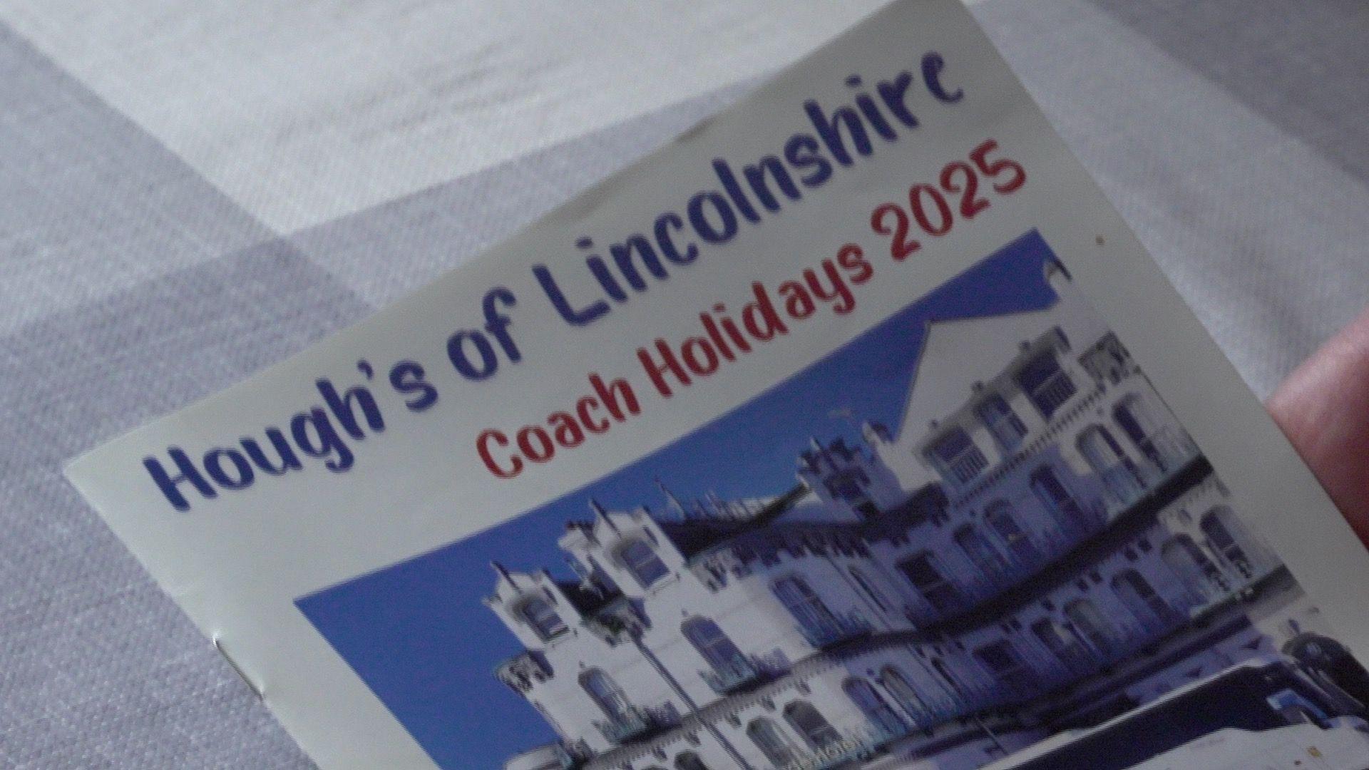 The front cover of a stapled holiday brochure held above a grey and white checked table cloth. The brochure is white and features an image of a large, white hotel beneath a blue sky. Blue text in a comic-sans-style font reads: "Hough's of Lincolnshire". Another line of text, in red, reads: "Coach Holidays 2025".