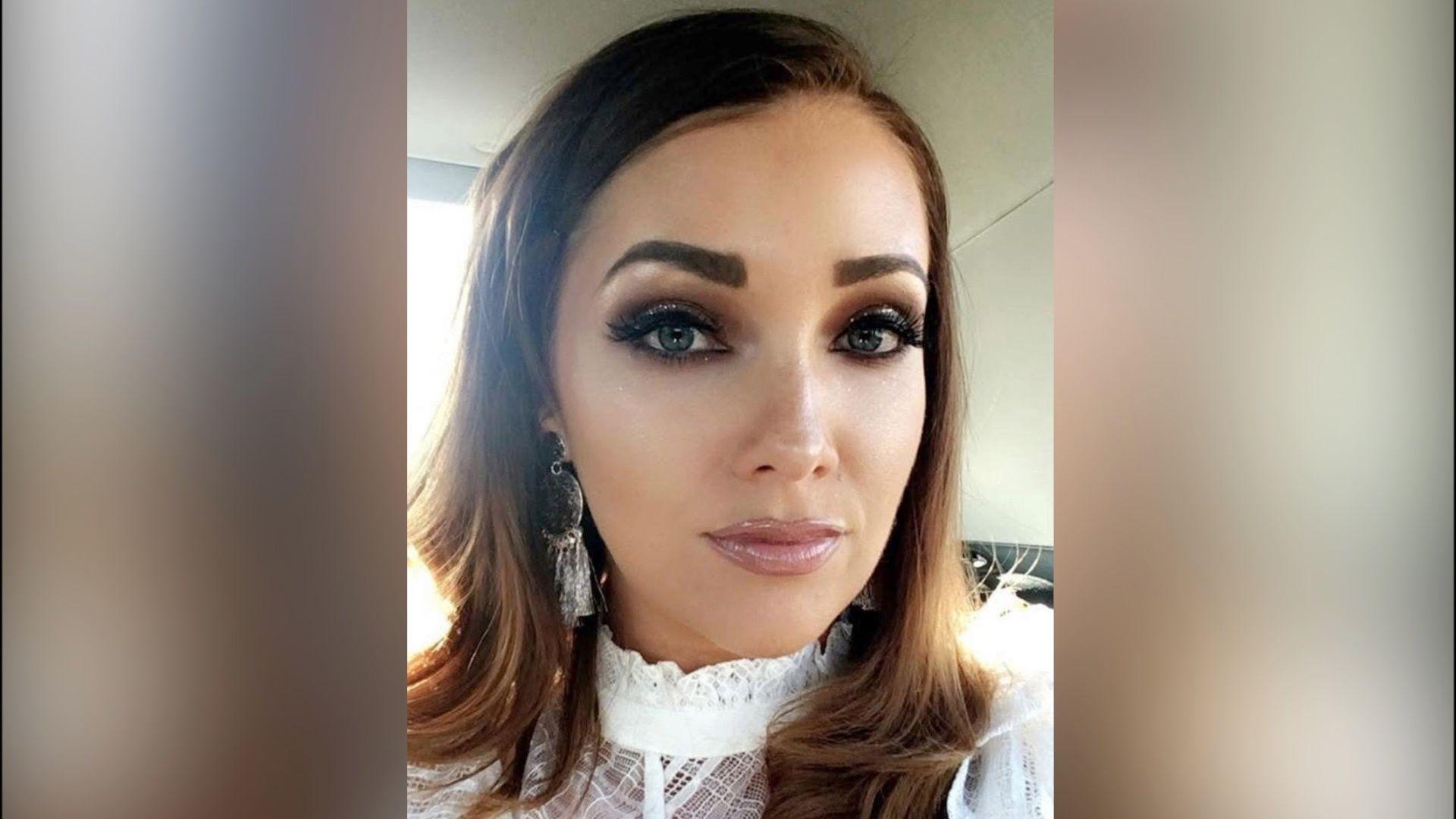 A selfie of a woman with shoulder length hair, dark eye make up and lip gloss. She wears a white lace top. 