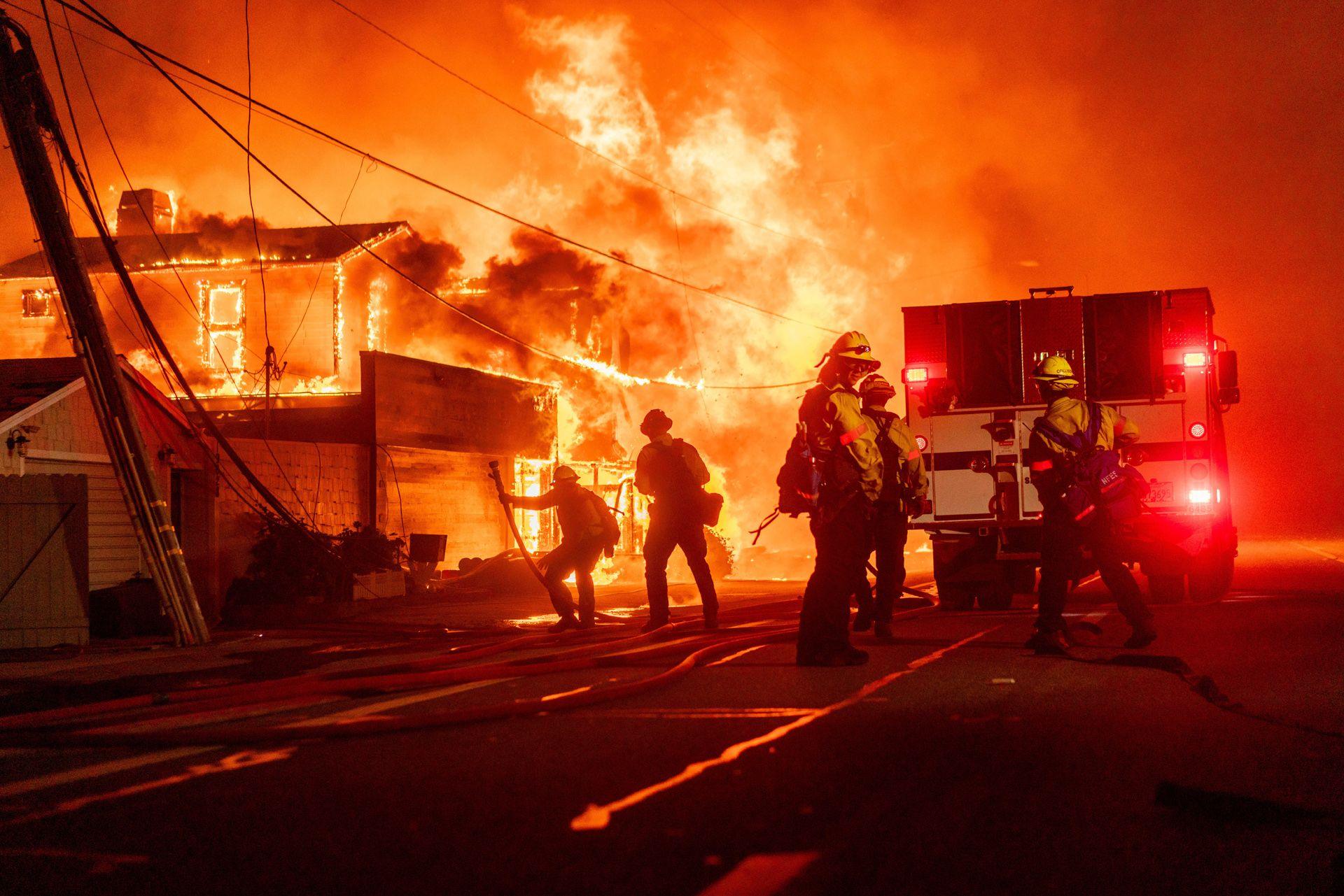 Silhouetted firefighters gather in front of a fire engine next to a burning house in the Pacific Palisades area