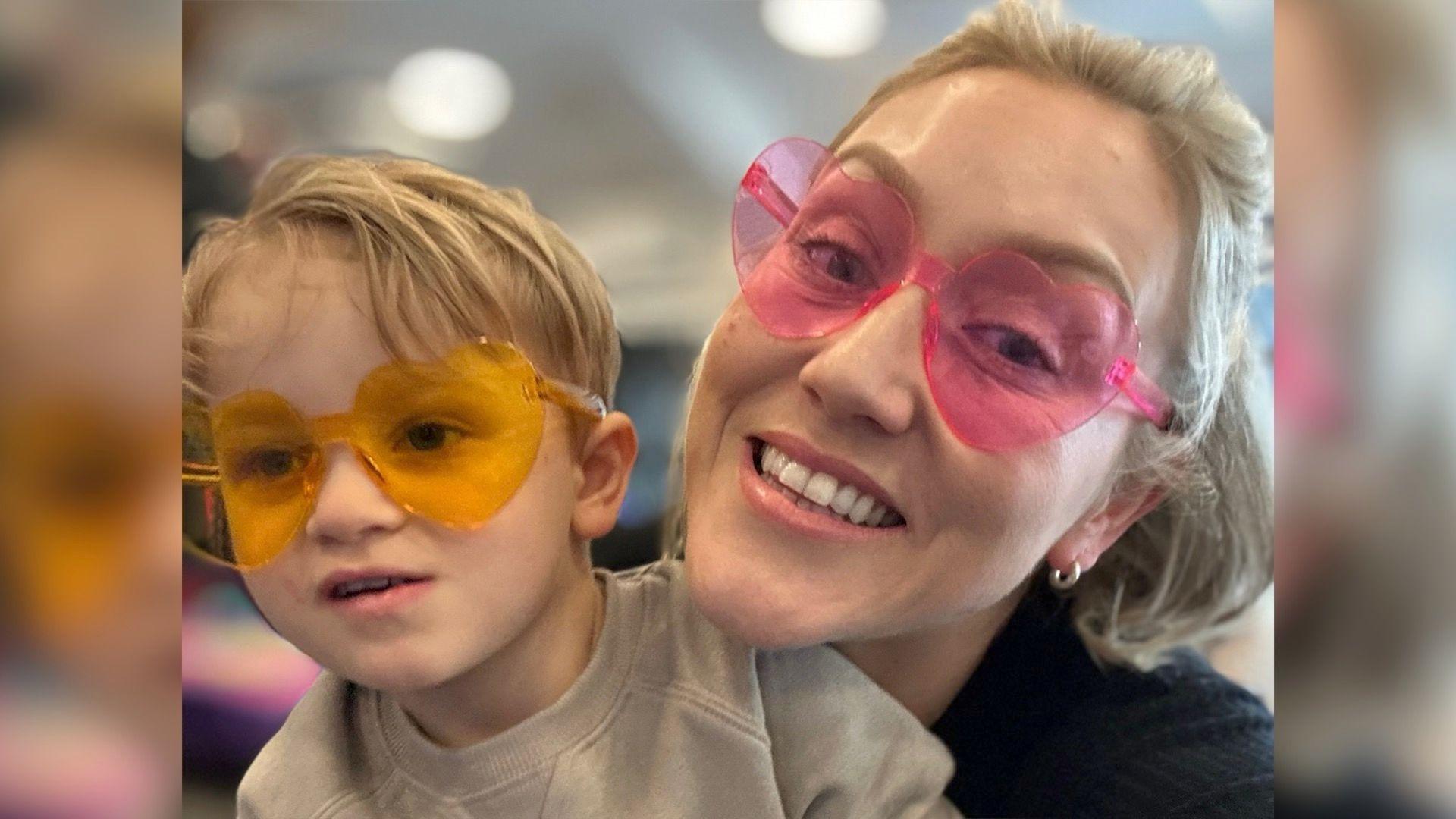 A small boy with blonde hair, wearing yellow love heart shaped glasses in held by his mother who also has blonde hair and is wearing pink ove heart shaped glasses.