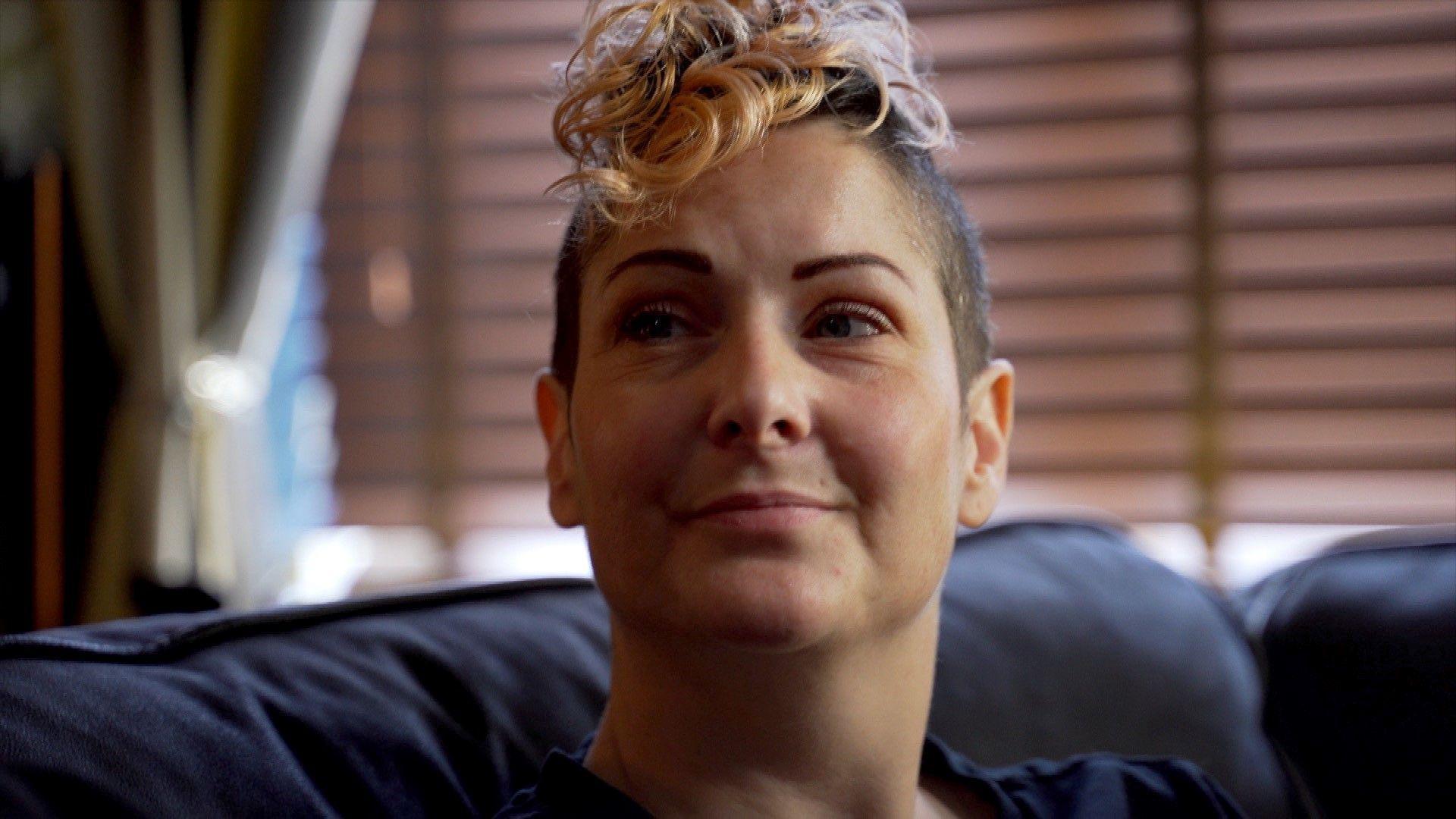 Clair Honeywood has short brown hair with a spiral of pink curls on top. She is seated on a blue sofa and looks to the side. 