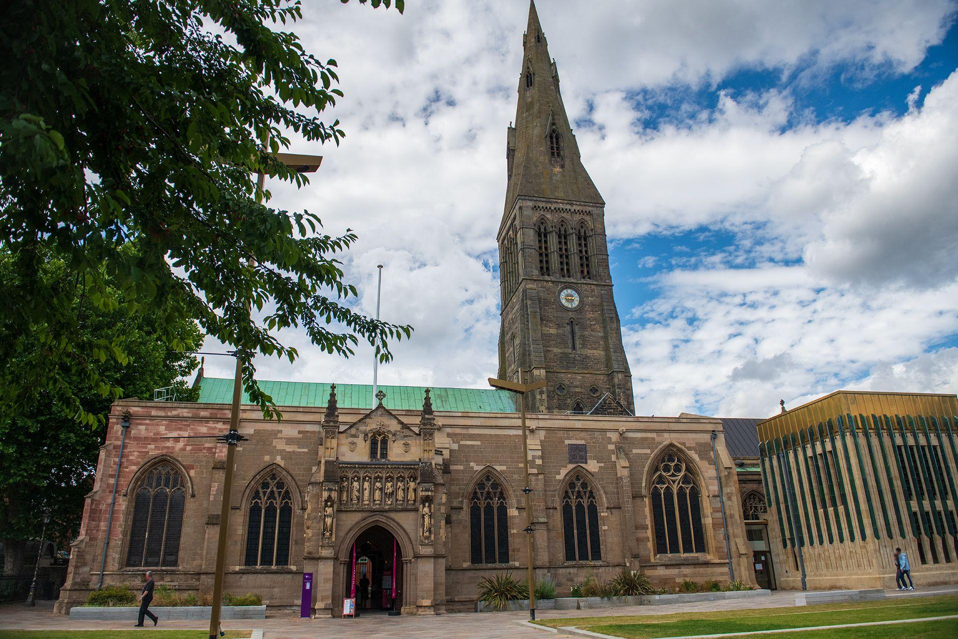 An exterior of Leicester Cathedral, towering high in front of a lawn with a large tree on the left.