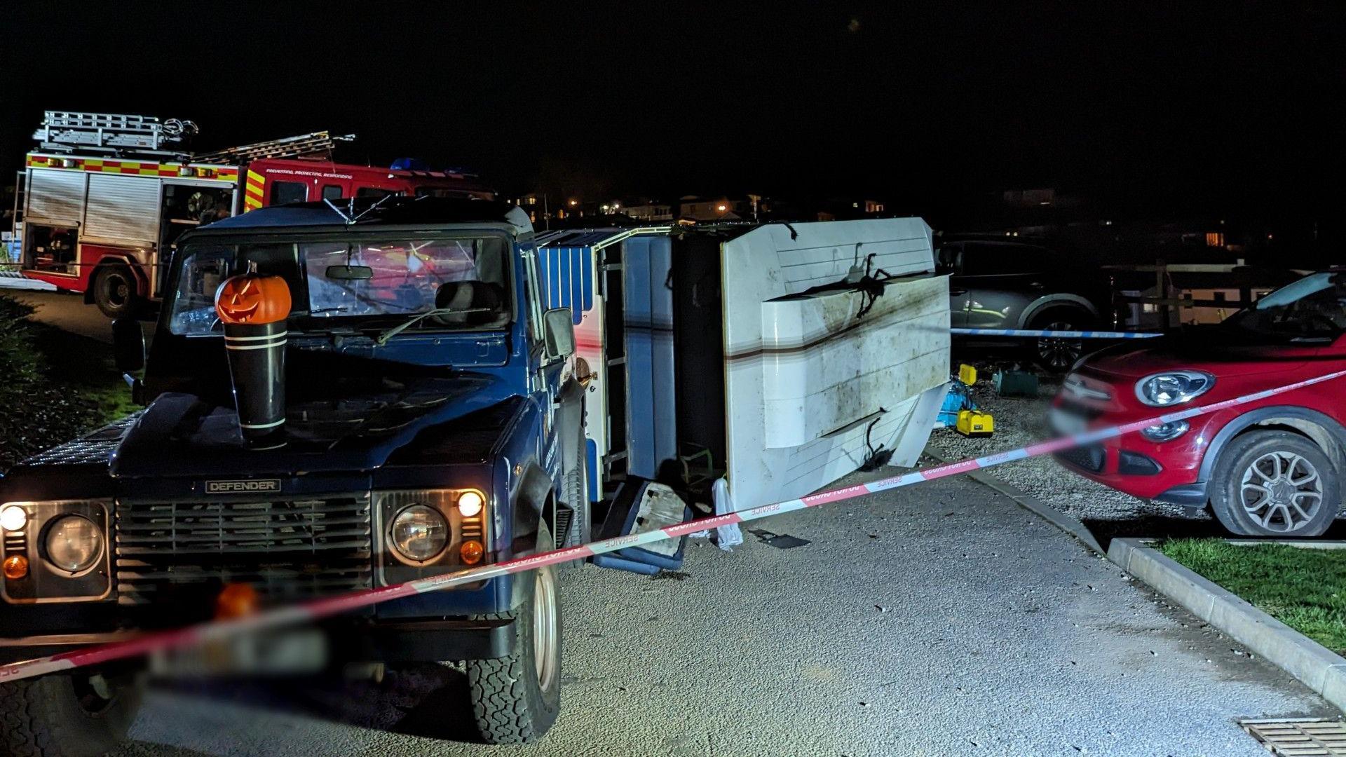 A Blue Land Rover Defender with a trailer that has tipped over at night. There is fire tape in the foreground and a fire engine in the background. 