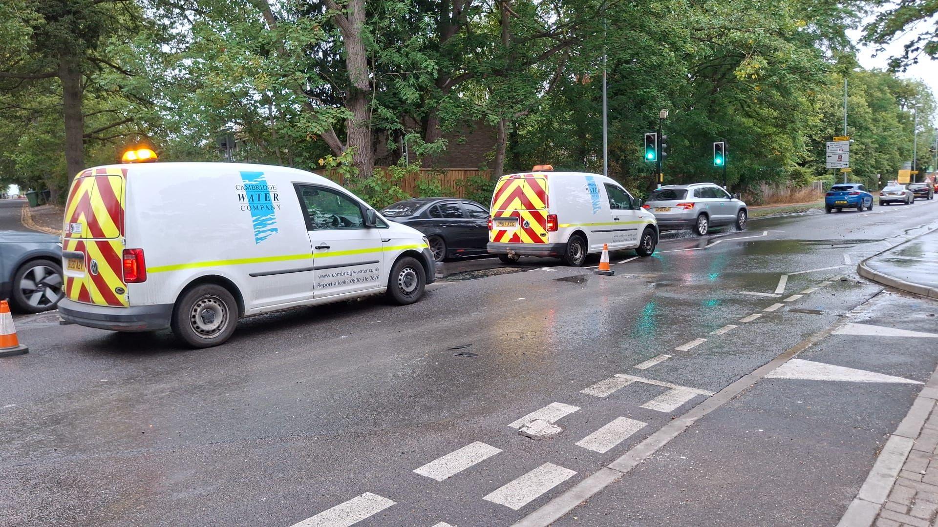 Water company vans are parked on a road which has a lot of water on it and cones sectioning off part of the carriageway. Cars are passing on the inside lane