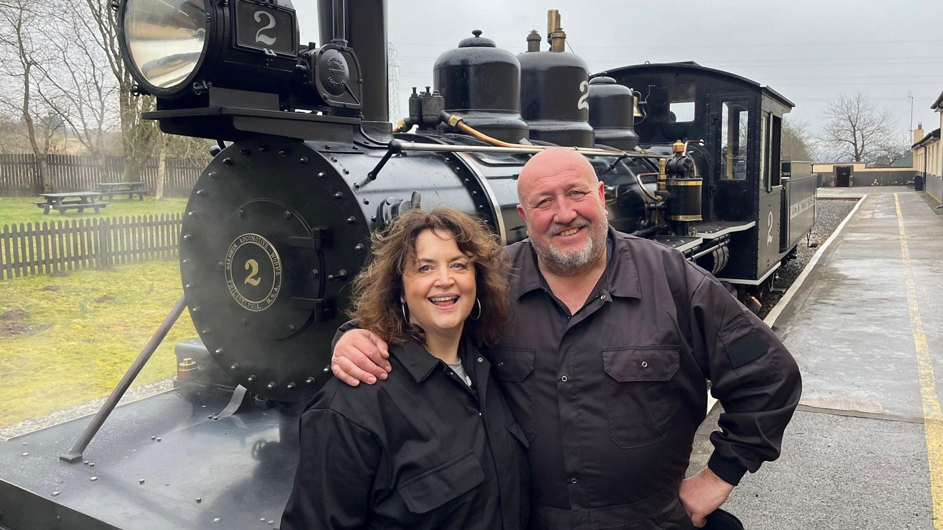 Ruth Jones and Steve Speirs share a hug on a railway platform with a black steam train in the background. They are both wearing black boiler suits.