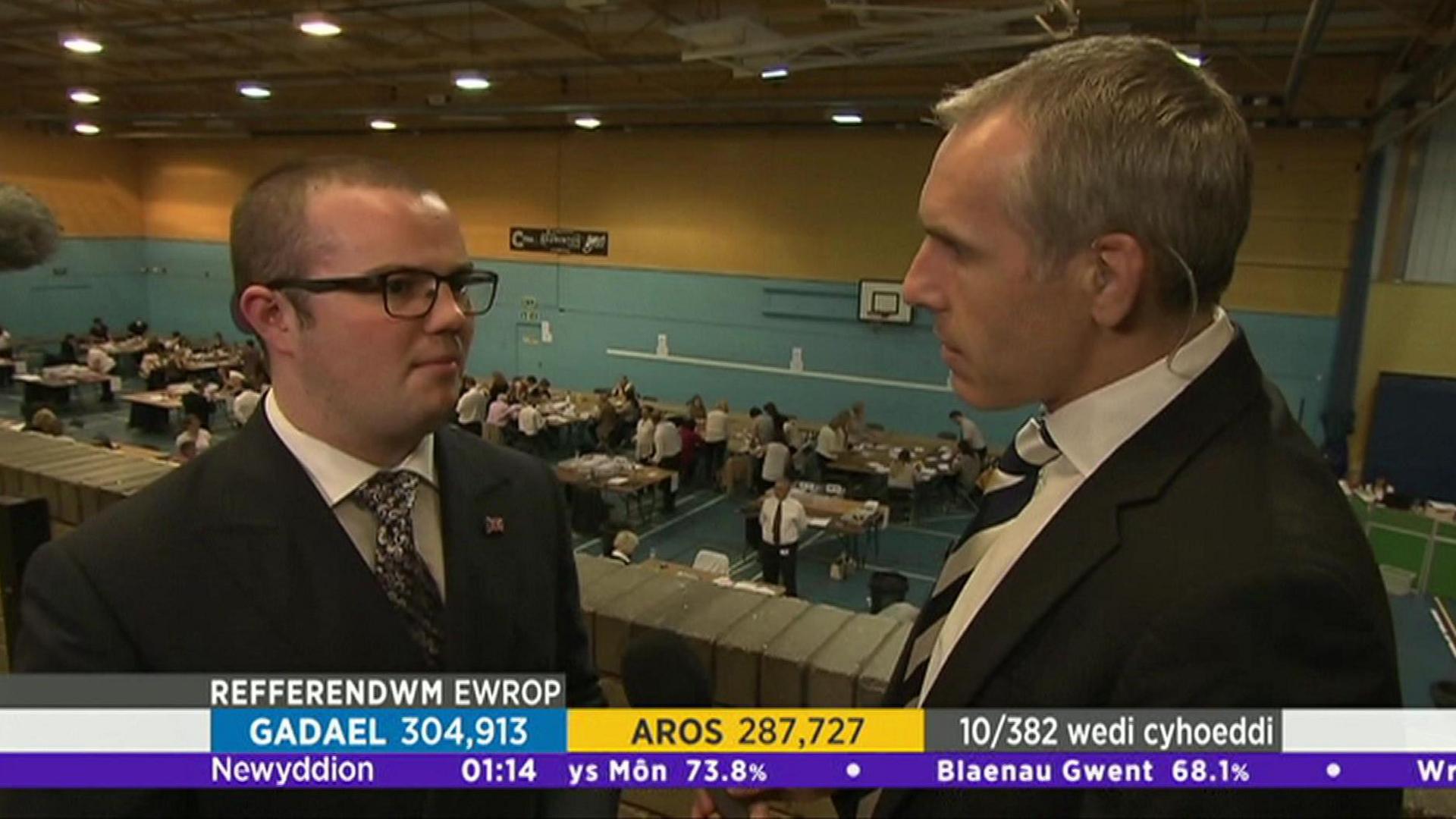 Mae'r llun yn dangos Llŷr Powell yn cael ei holi gan ohebydd.  Mae'r ddau'n sefyll uwchlaw neuadd chwaraeon ble mae pleidlieisiau yn cael eu cyfrif.