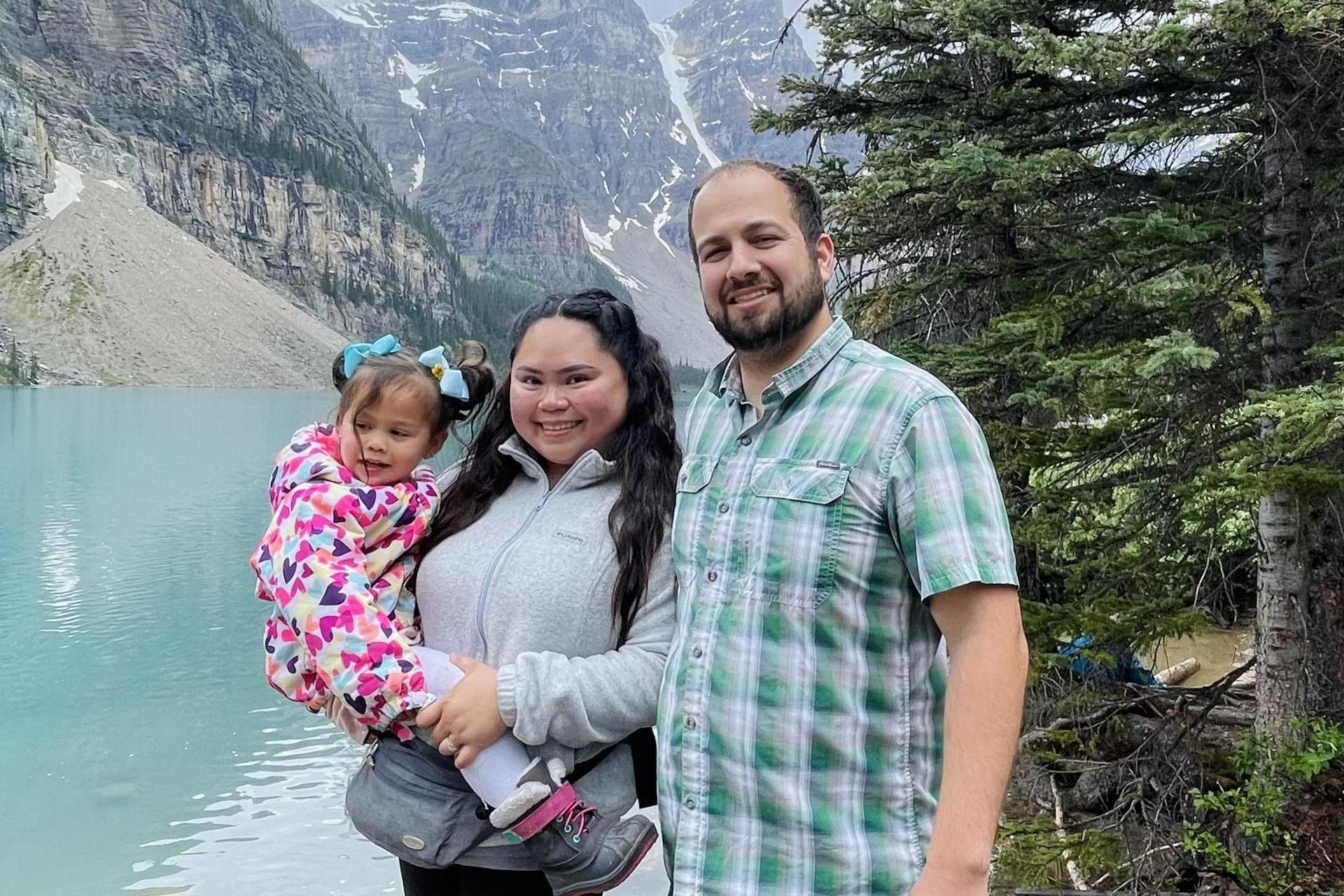A man stands next to his wife as she holds their daughter on her hip. A lake with mountainous sides can be seen in the background, as well as a conifer tree.