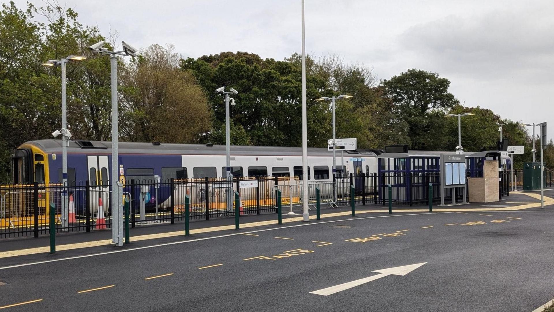 Ashington station with a two carriage train waiting at the platform. There are black railings between the platform and a large carpark 