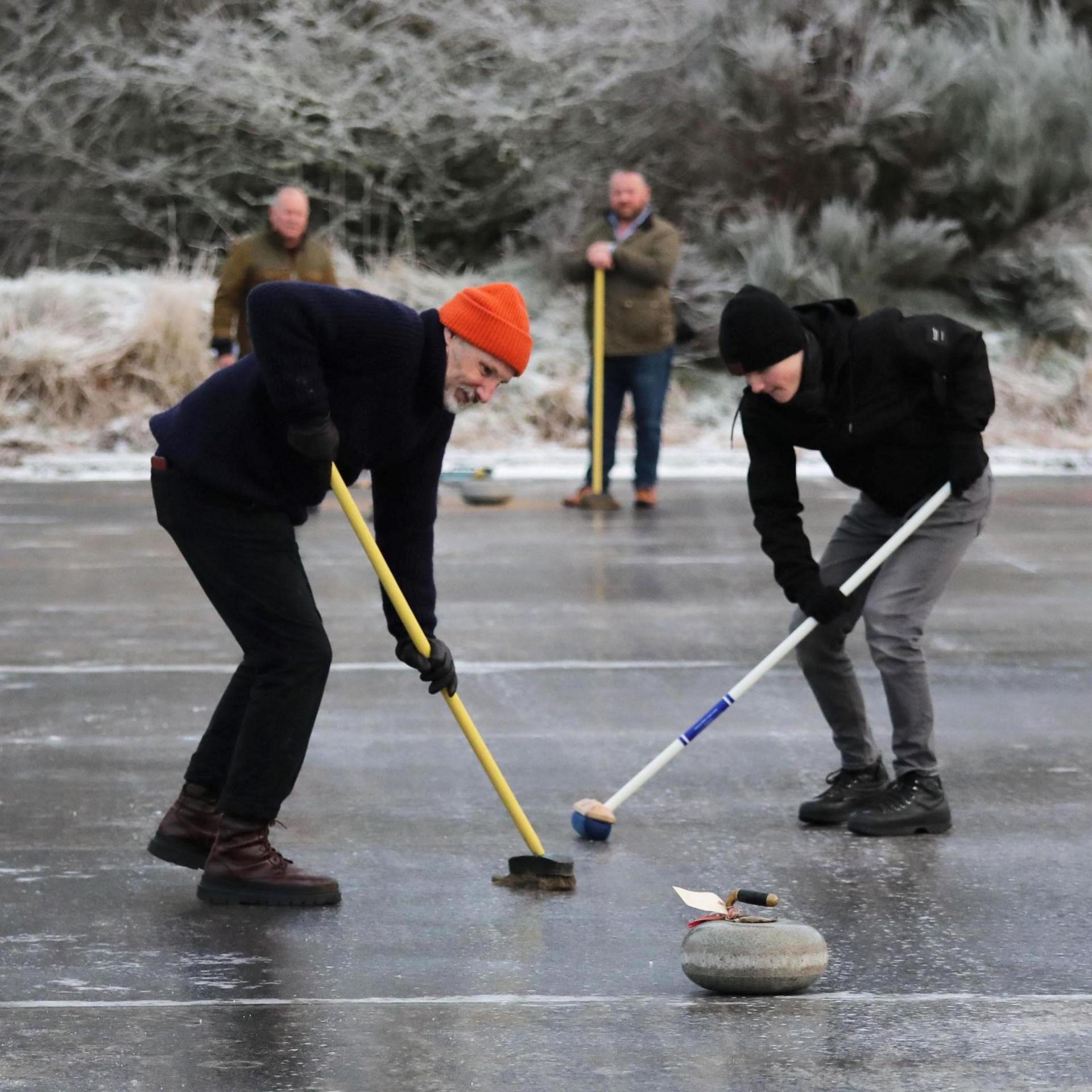 Two curler use brushes to brush the ice to help a curling stone travel down the rink. The men are wearing hats and gloves. Two other curlers watch from the sidelines.