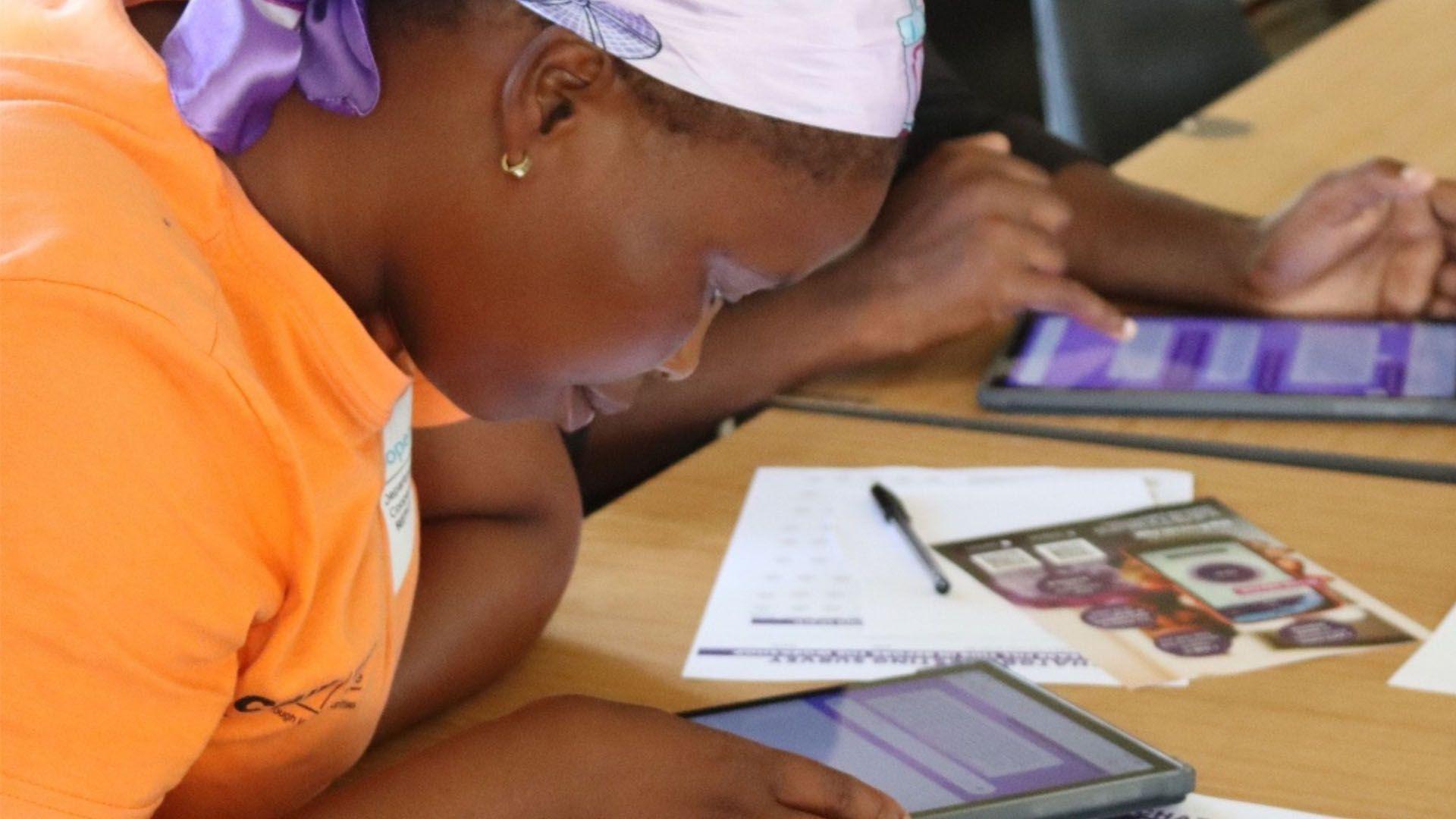 A woman sits at a table using a tablet during a digital training session. She is wearing an orange shirt and a patterned headscarf. In front of her are a pamphlet about using technology to stop gender-based violence, and other learning materials.