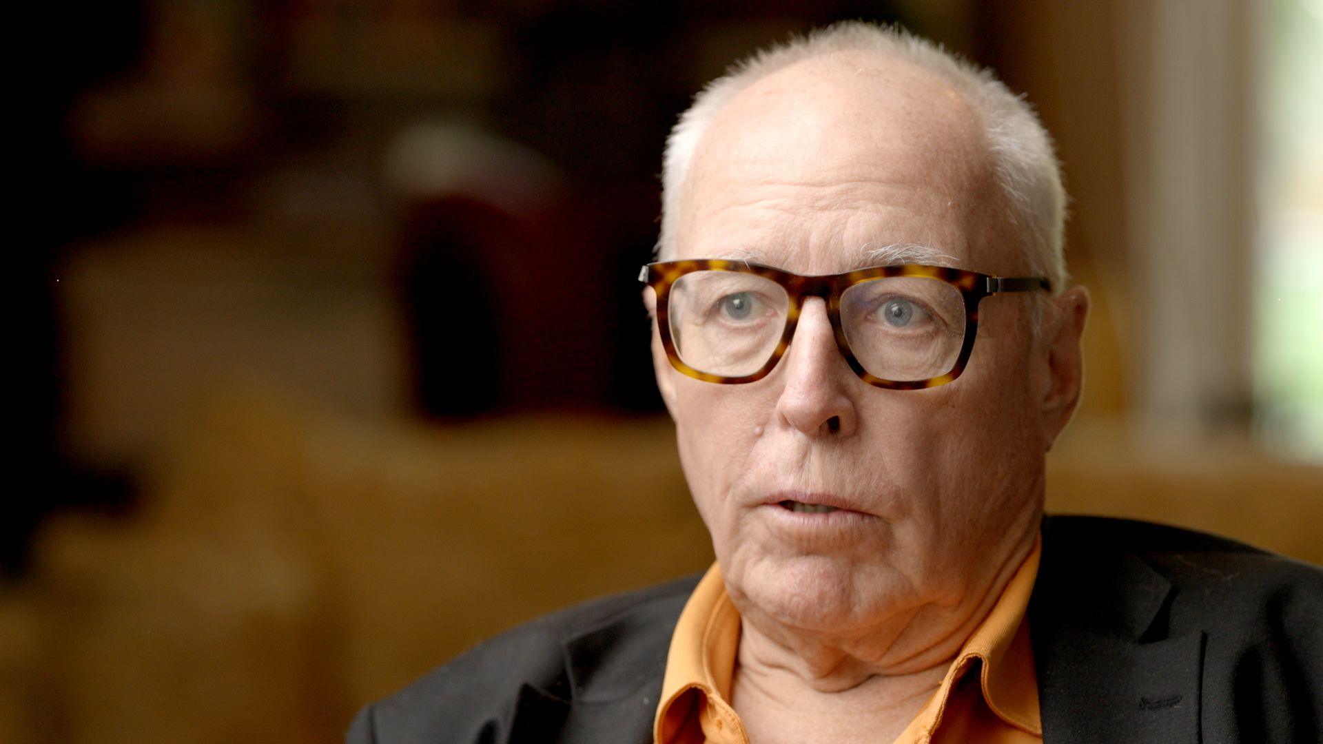 Mark Schneider, pictured in close-up as he is being interviewed, with the background blurred. He has white hair, clipped short, and wears glasses with a tortoiseshell-style frame, an orange shirt and a black jacket.