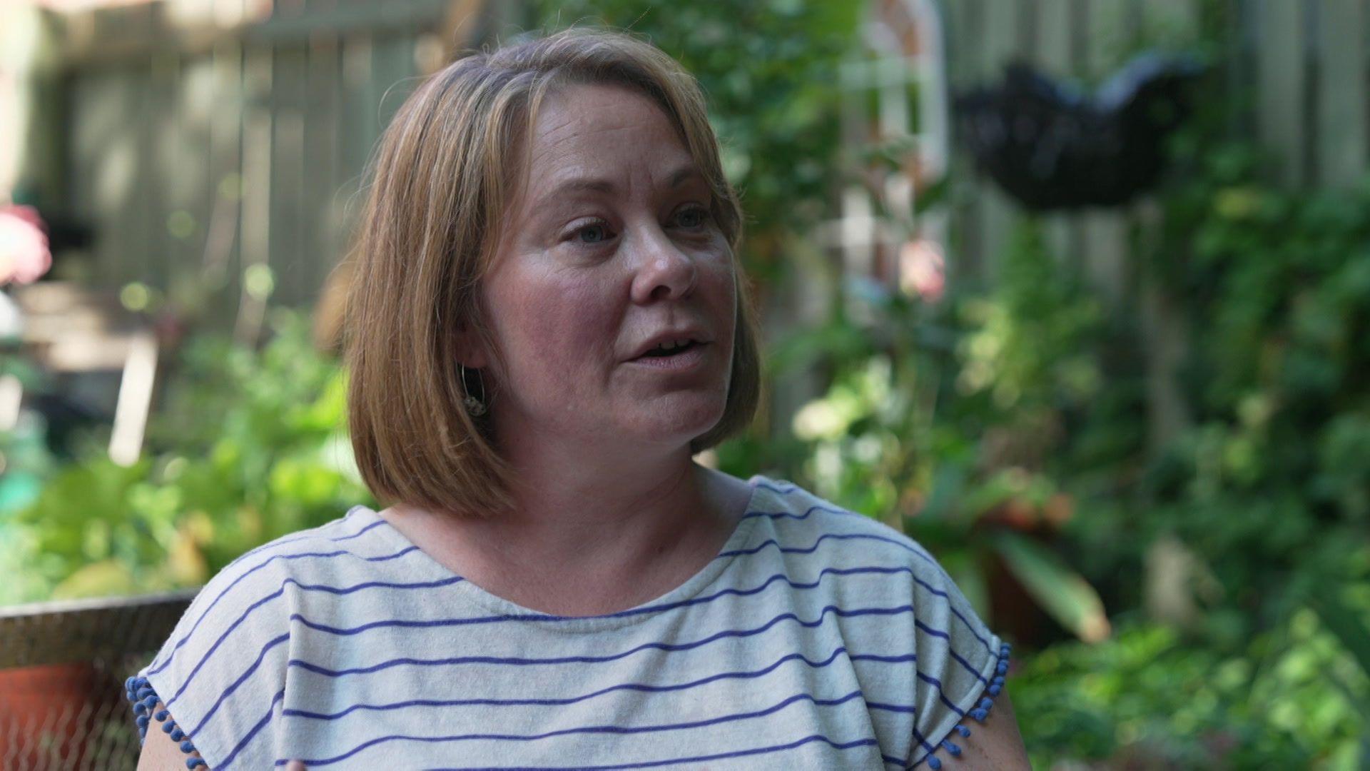 Dr Megan Klaar, a woman with shoulder-length fair hair and a striped t-shirt, sits in a garden. Blurred plants can be seen behind her.