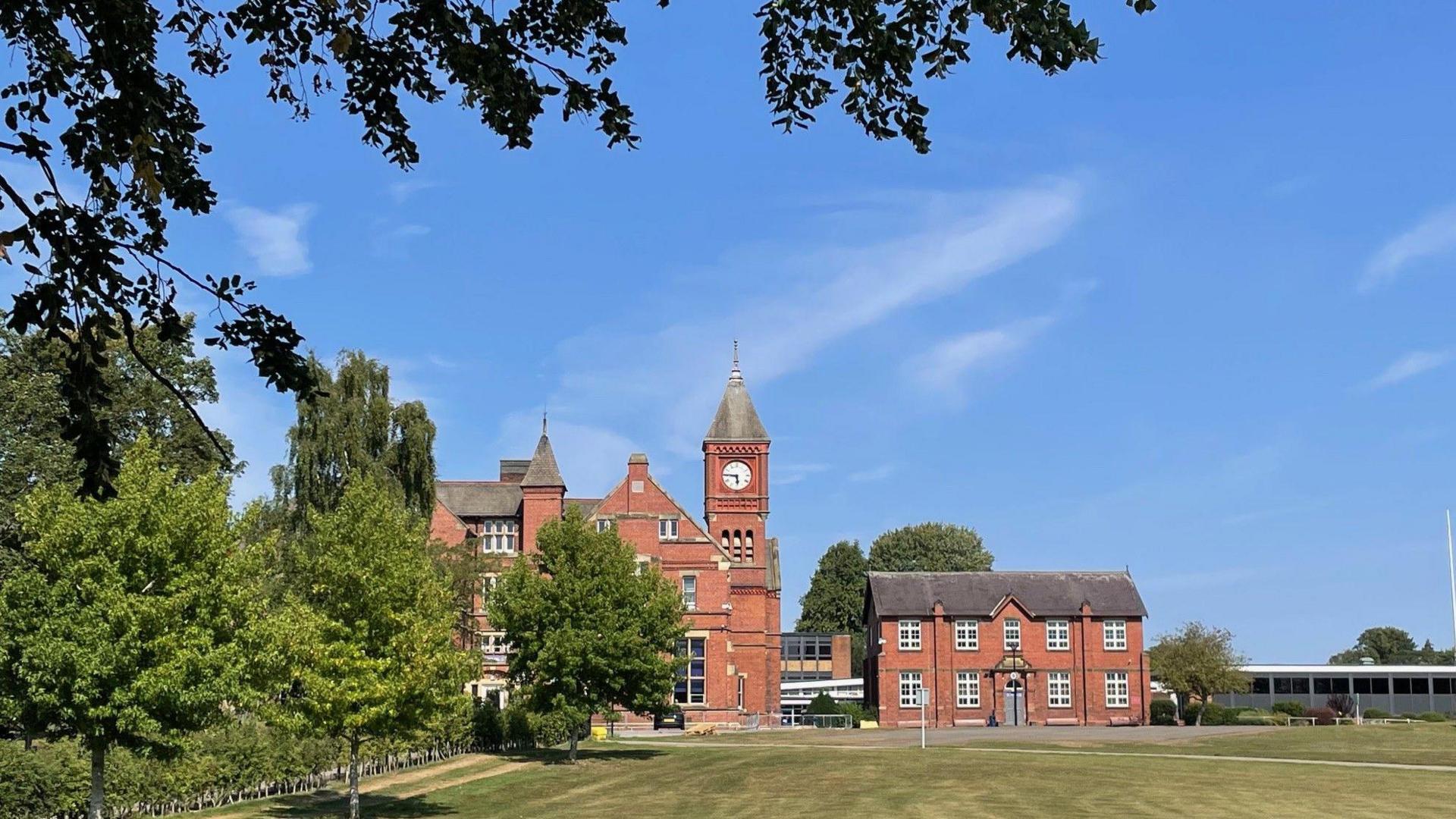A red brick school with a clocktower and a green lawn in front and a bright blue sky.