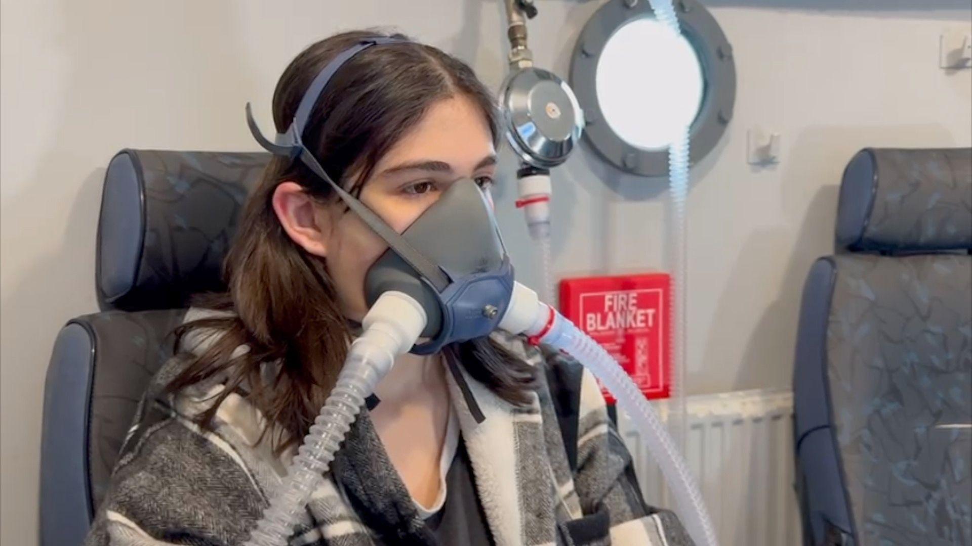 A girl with dark hair is sat on a chair and has an oxygen mask over her nose and mouth.