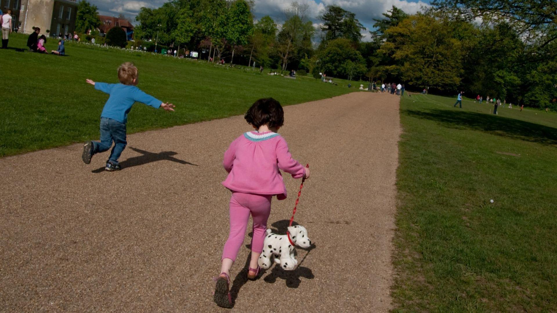 Two children run along a path in Priory Park, Reigate. The boy has his arms outstretched in a flying position and the girl is wearing a pink suit and has a toy pet