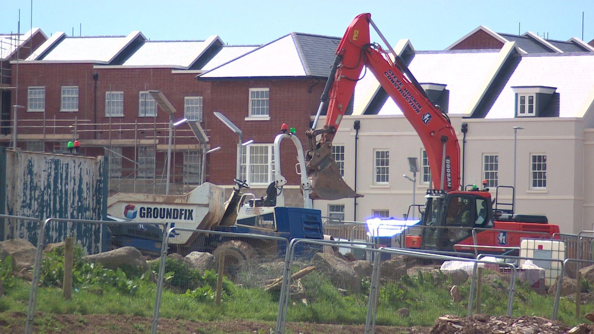 A red digger and a white truck working on a fenced construction site. There are multiple new looking residential homes with some scaffolding in place.