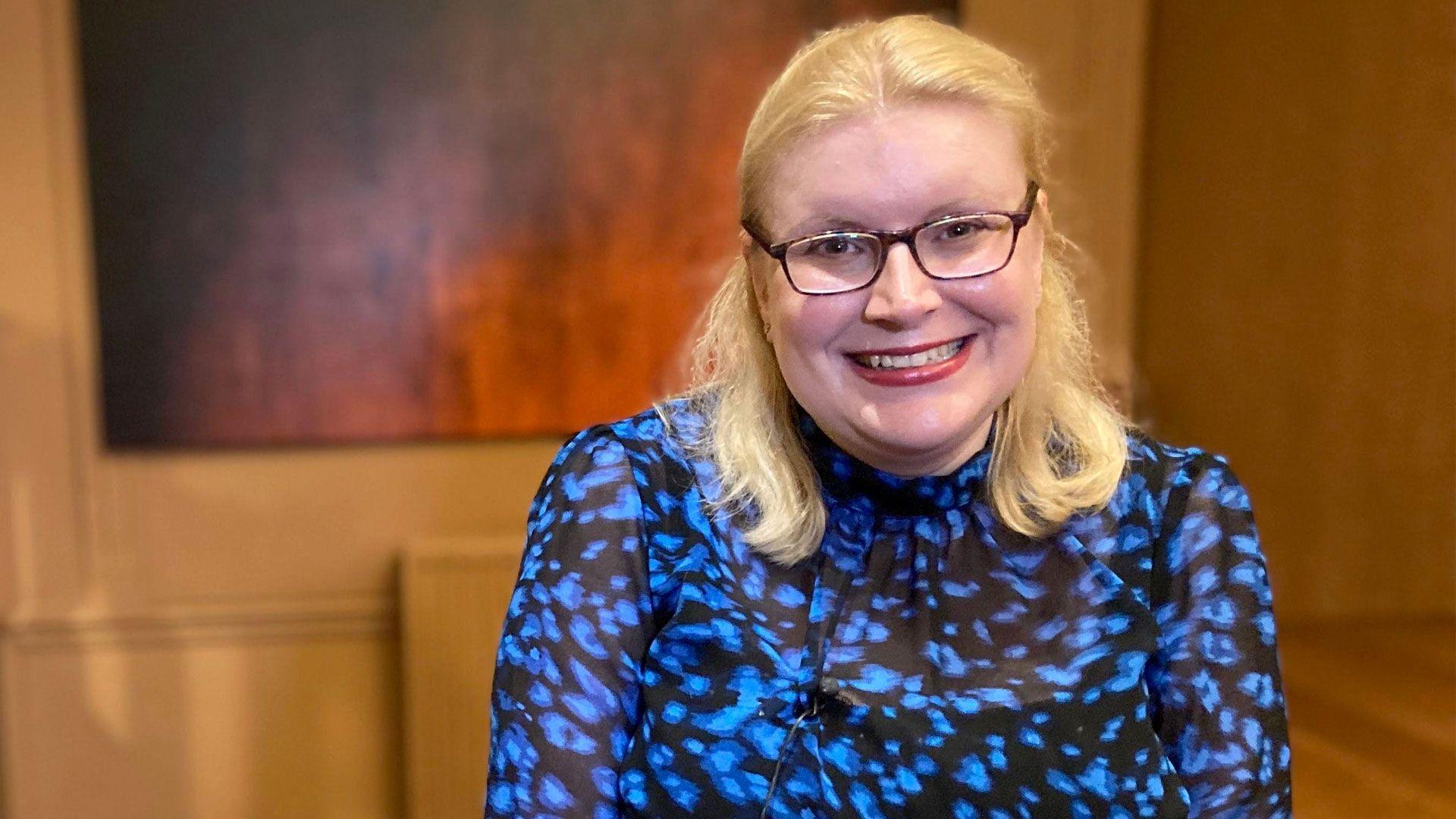 Laura with blonde hair and glasses smiling as she looks into the camera. She has a blue and black patterned top on and is sitting in the corner of a room with a large picture on a wall behind her.